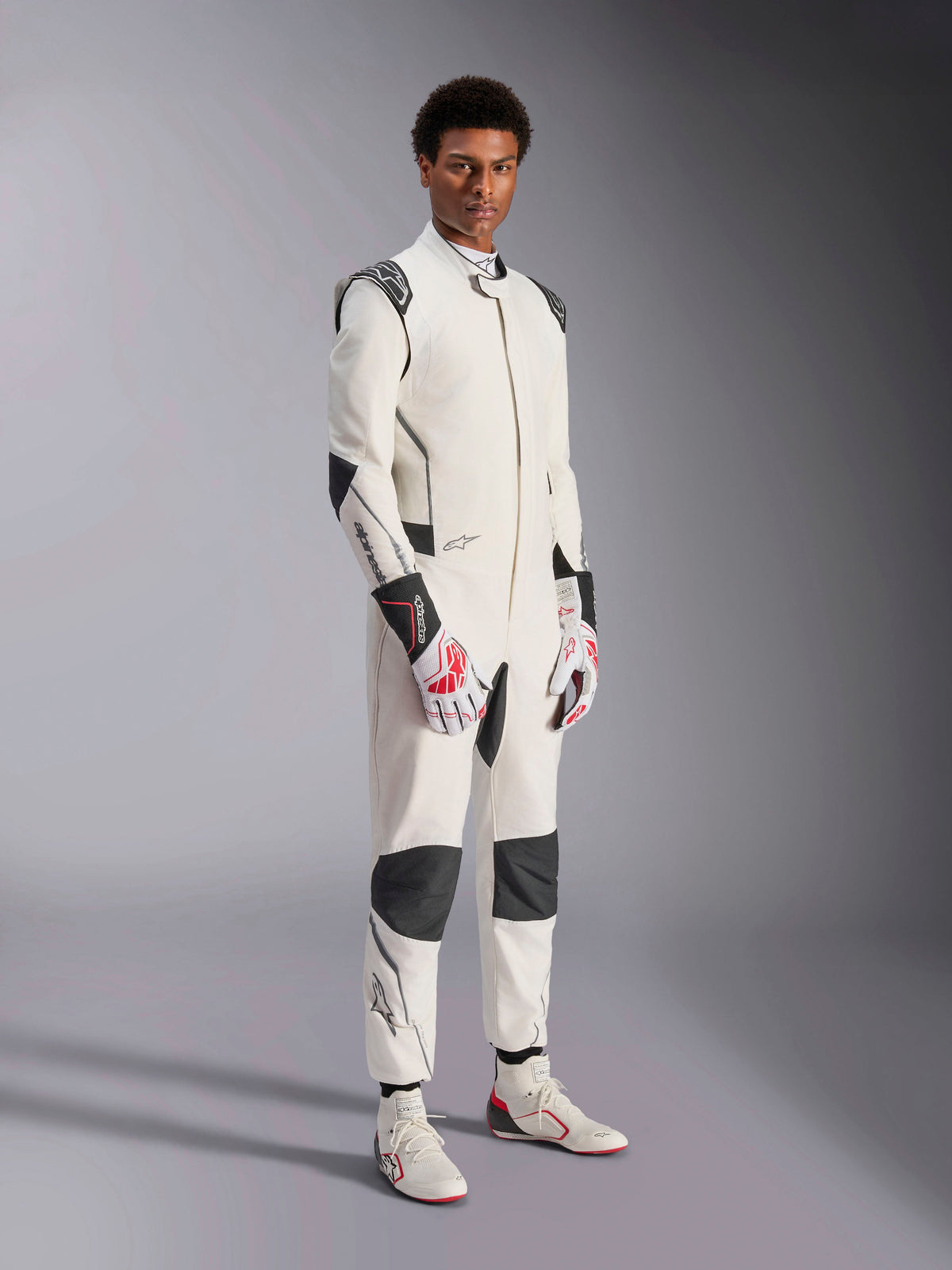 Manlig modell i Alpinestars Hypertech V3 Suit, vit med svarta och silvriga detaljer, professionell bilracingutrustning med svarta paneler på axlar och knän, matchad med vita och röda racinghandskar och vita racingstövlar