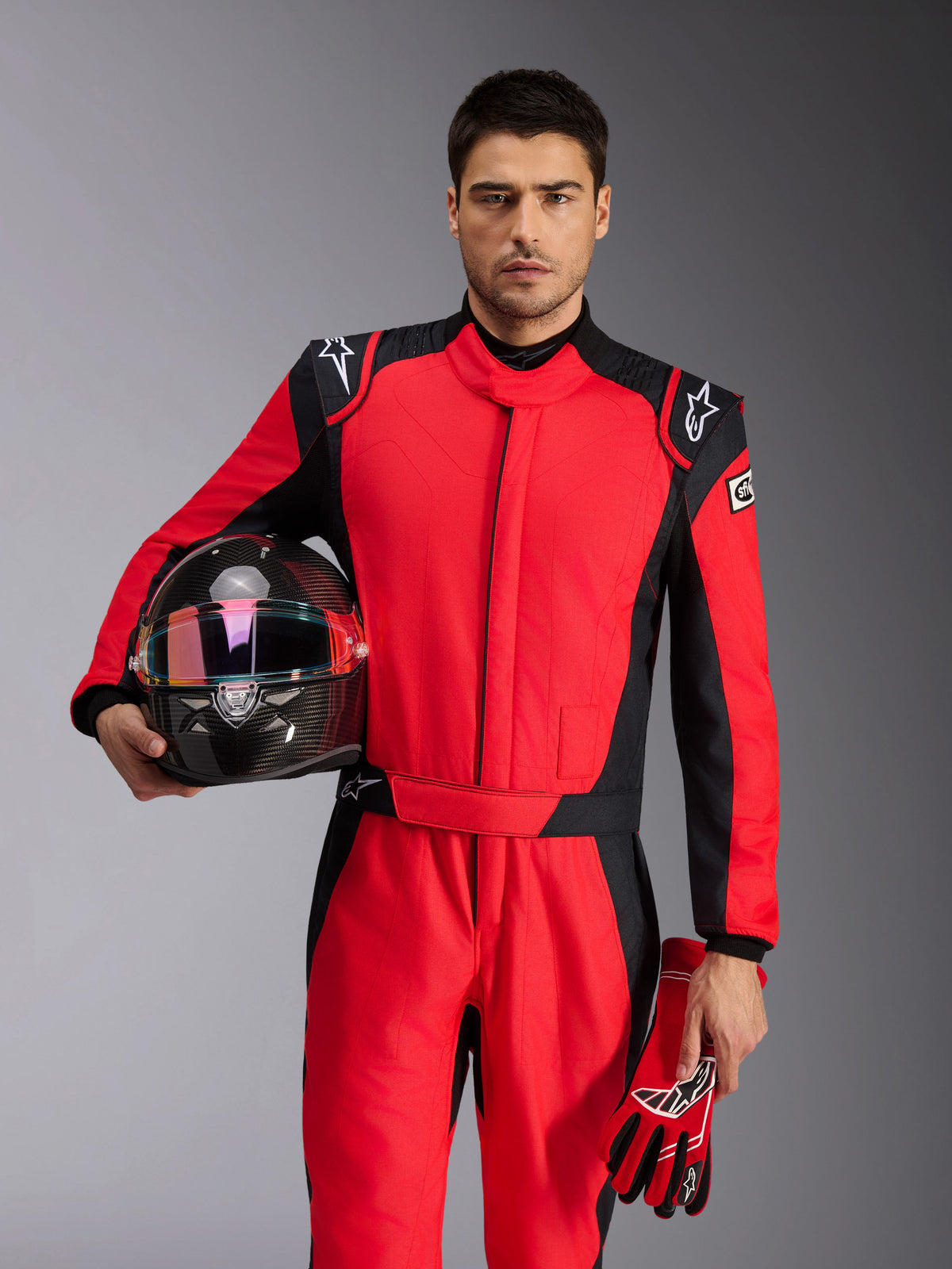 Manlig racerförare bär Alpinestars GP Pro Comp V2 FIA-overall i rött och svart, håller en racinghjälm i kolfiber och matchande handskar, professionell motorsportklädsel