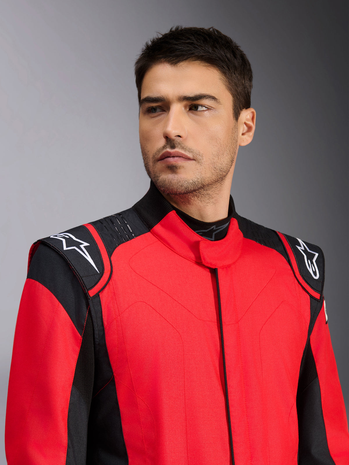 Manlig modell iklädd Alpinestars GP Pro Comp V2 FIA racingoverall, röd och svart, med vita Astars-logotyper på axlarna och detaljer med silikongreppsteknik, professionell motorsportutrustning