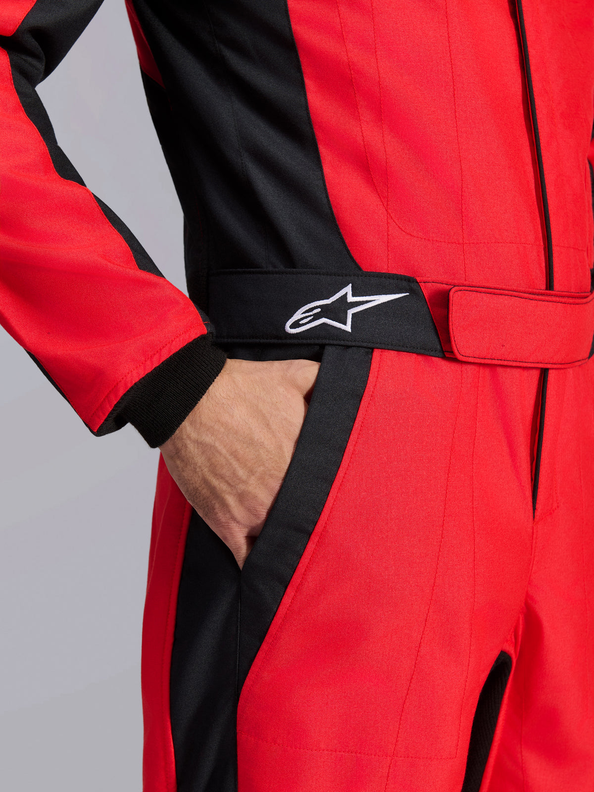Alpinestars GP Pro Comp V2 FIA racingoverall, röd och svart, närbild av midjepartiet som visar en hand i fickan, med ett svart bälte med en vit stjärnlogotyp och strukturerade paneler för bilsport