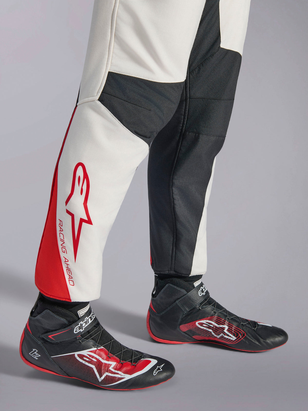Alpinestars GP Tech V4-overall, racingoverall, vit, svart och röd, vy över benens nederdel med vita och svarta paneler och röda detaljer samt 'RACING AHEAD'-märkning, buren med svarta racingskor, professionell motorsportutrustning