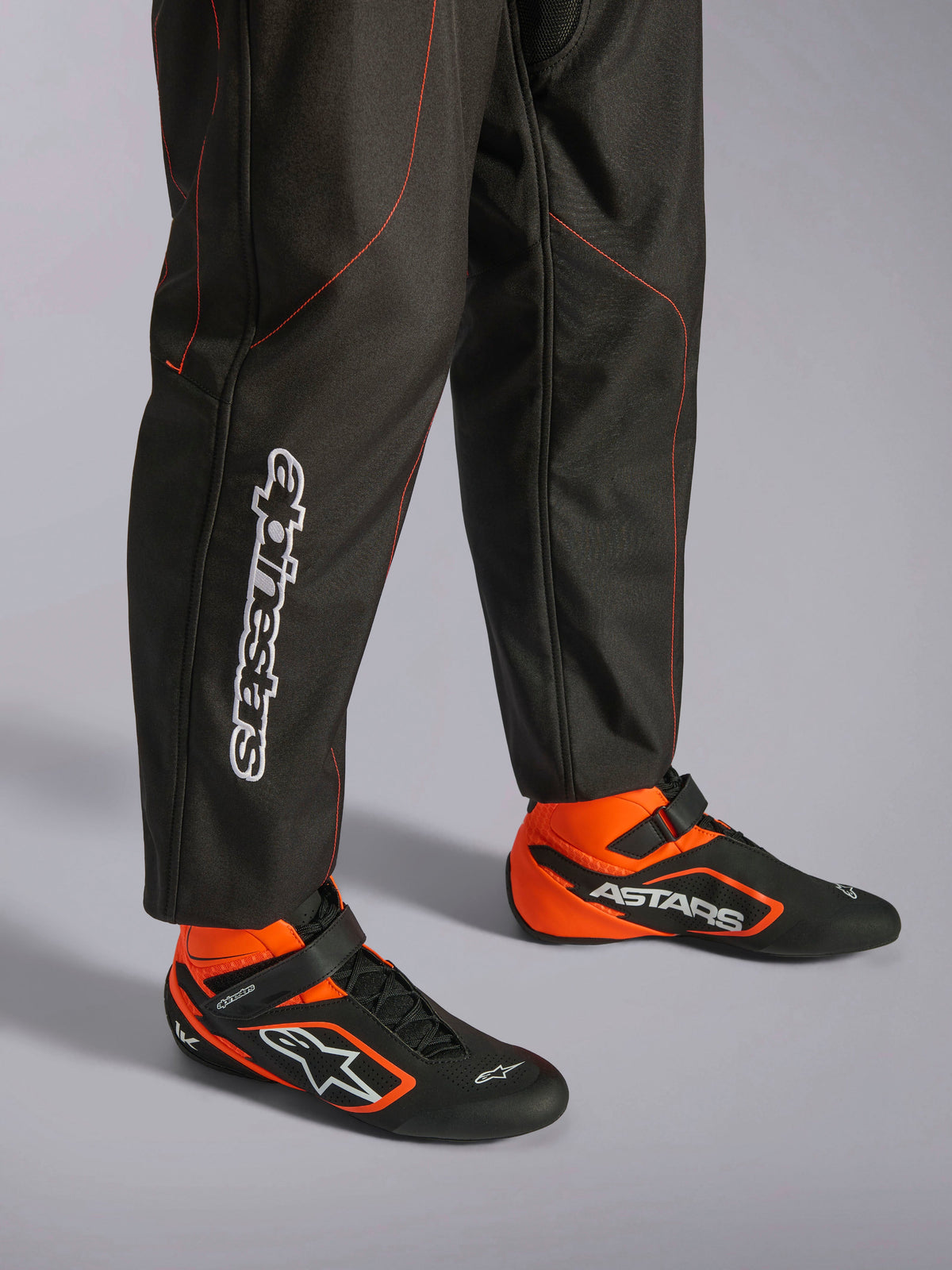 Alpinestars KMX-9 V3 Suit, Karting Suit, Black and Red Fluo, detailed view of the lower legs and racing shoes, black fabric with fluo red stitching, vertical Alpinestars logo on the calf, paired with matching black and orange racing boots, designed for professional karting