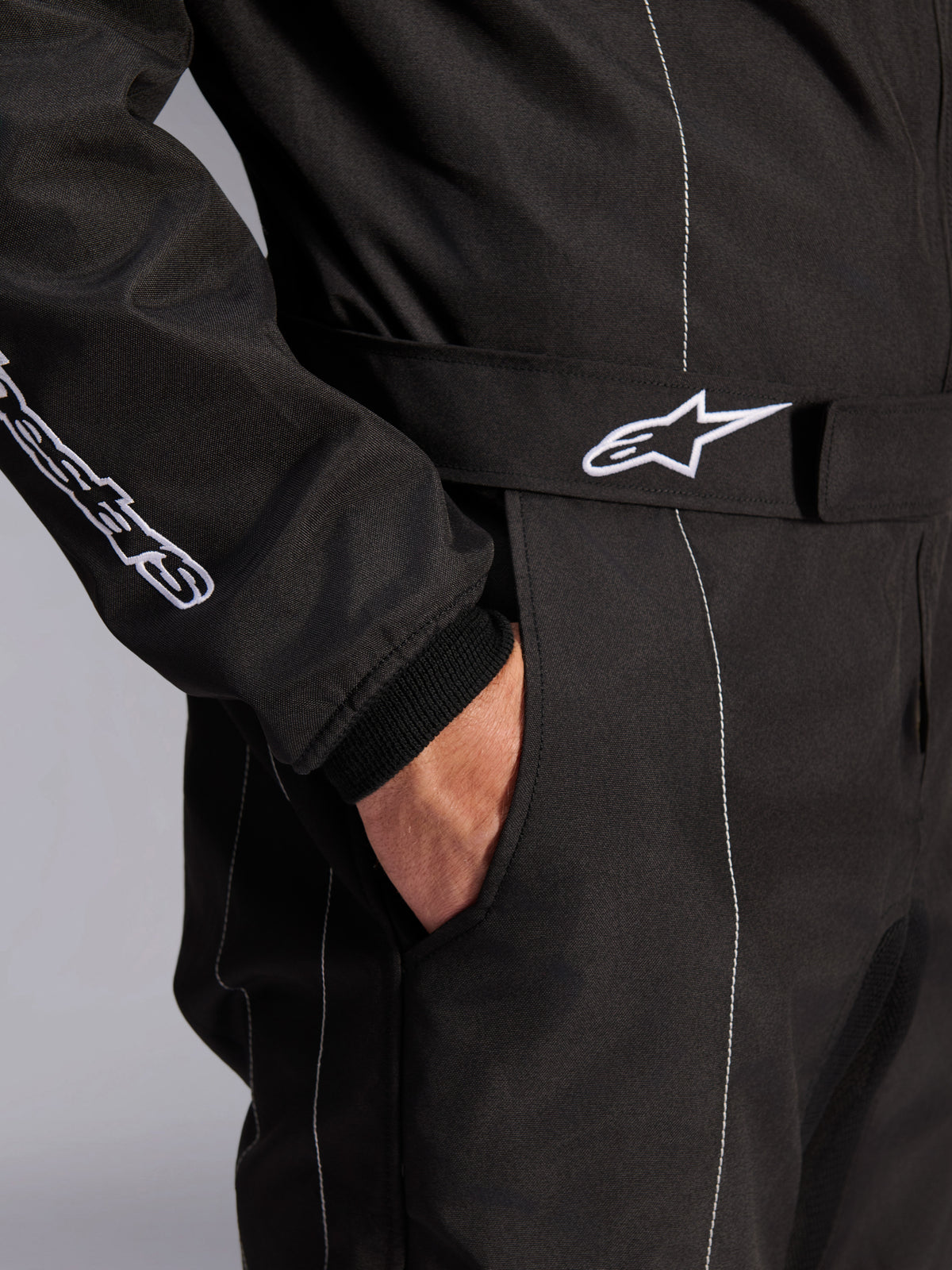 Alpinestars KMX-9 V3-dräkt, svart och vit, närbild på justerbart midjebälte med broderad logotyp, sidoficka, vita kontrastsömmar på svart tyg, teknisk detalj på kartingdräkt