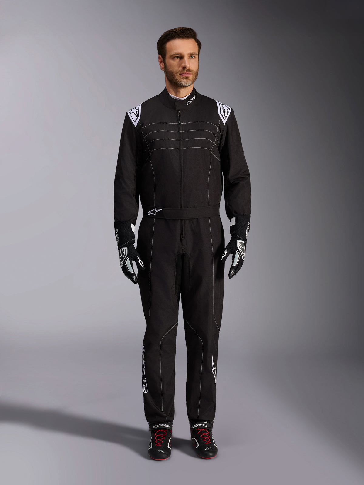 Alpinestars KMX-9 V3-ställ, svart och vit kartingoverall, helfigur på en modell, med vita axellogotyper och detaljer, stylad med svarta och vita racinghandskar och svarta skor med röda skosnören, athletic-regular passform för professionell kartingutrustning.