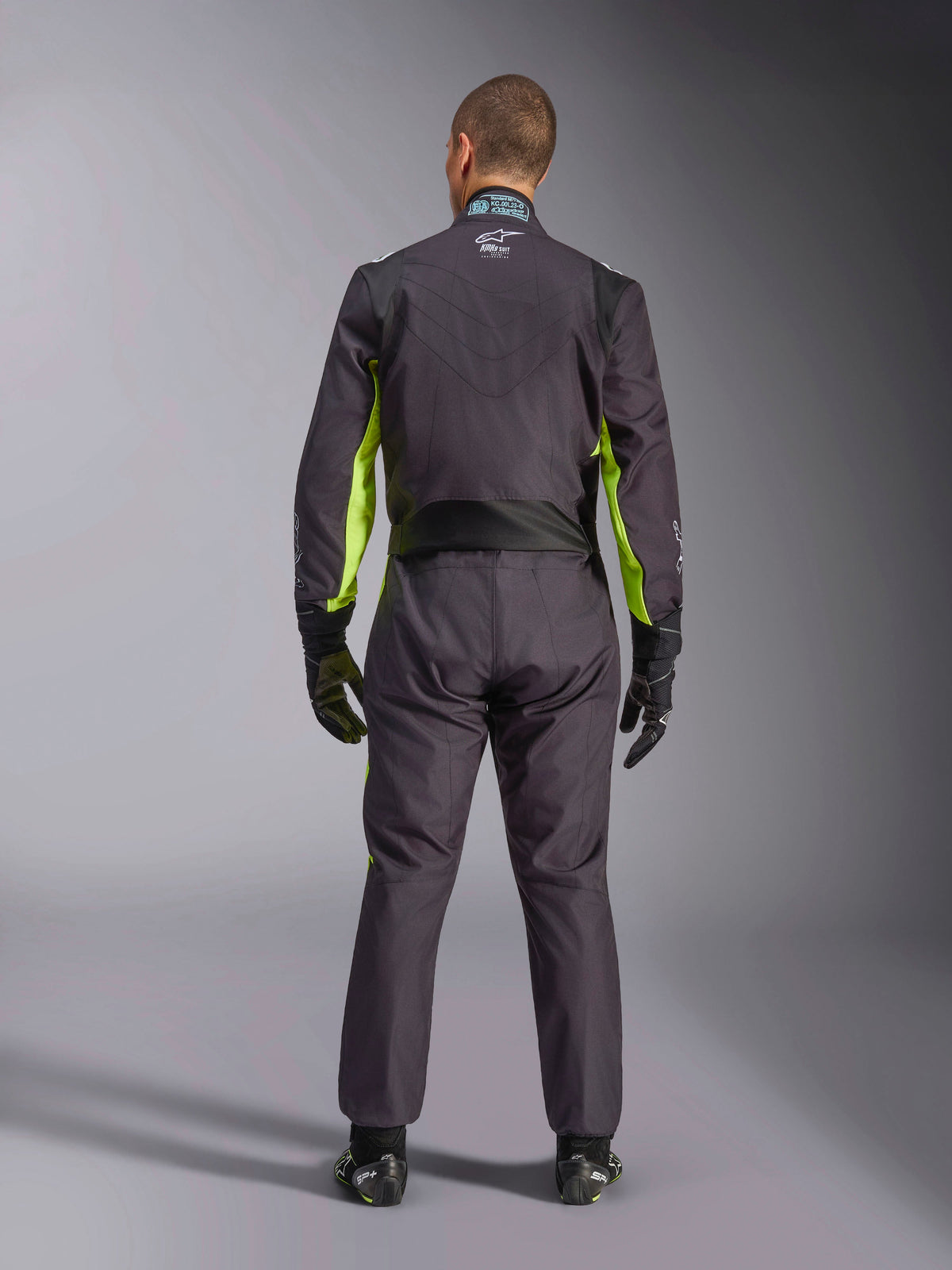Full-body rear view of a person wearing the Alpinestars KMX-9 V3 Graphic 3 Suit, Karting Suit in Black and Yellow Fluo, showing ergonomic stretch panels on the back and shoulders, paired with black racing gloves and footwear.