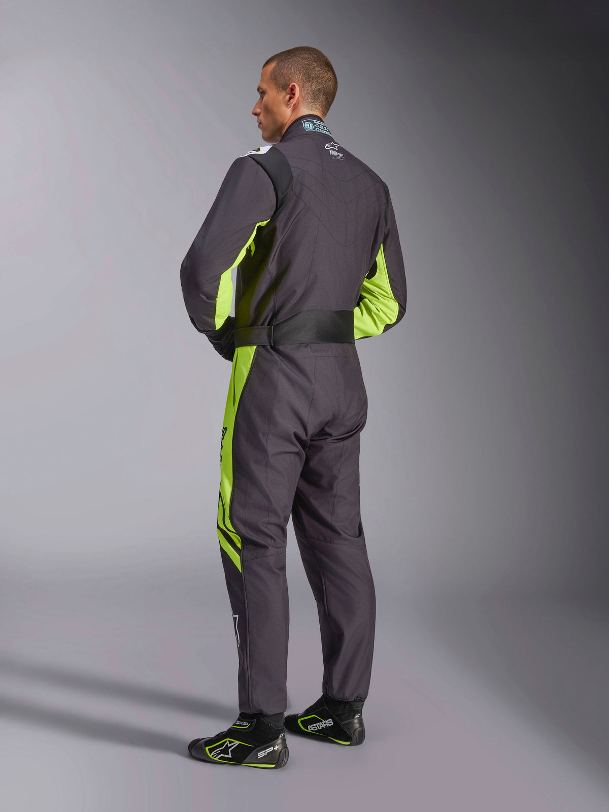 Alpinestars KMX-9 V3 Graphic 3 Suit, Karting Suit, Black and Yellow Fluo, back view of model showing the one-piece design with fluorescent yellow accents on the sides and underarms, ergonomic stretch panels on the lumbar area, and adjustable waist belt, worn with coordinating racing shoes.
