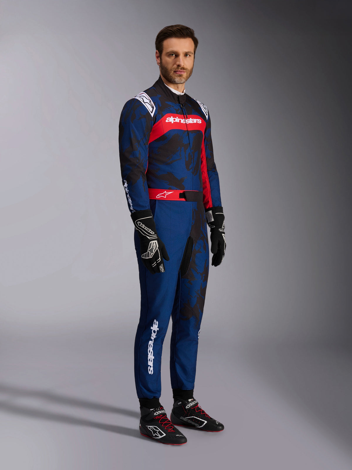 Modell som bär Alpinestars KMX-9 V3 Graphic 5 Suit i Navy Red, helkroppsbild inklusive svarta kartinghandskar och svarta racingskor, marinblå dräkt med mörk grafik i kamouflagestil och en framträdande röd bröstpanel med vit logotyp, professionell skyddsutrustning för karting