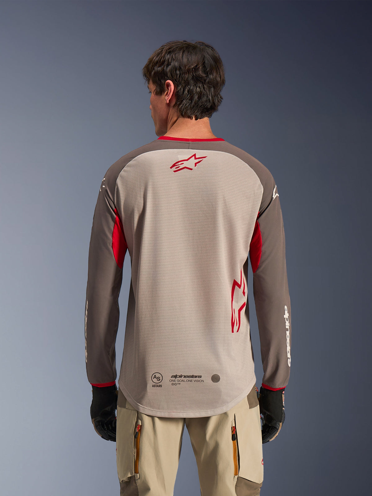 Baksida av en man som bär Alpinestars Maxdura Dual Jersey i Aluminum Walnut, en äventyrströja för motorcykel med beigefärgad meshtorso och röd stjärnlogotyp, mörkbruna ärmar med röda detaljer, matchad med beige körbyxor och svarta handskar, designad för högpresterande ventilation vid terrängkörning.