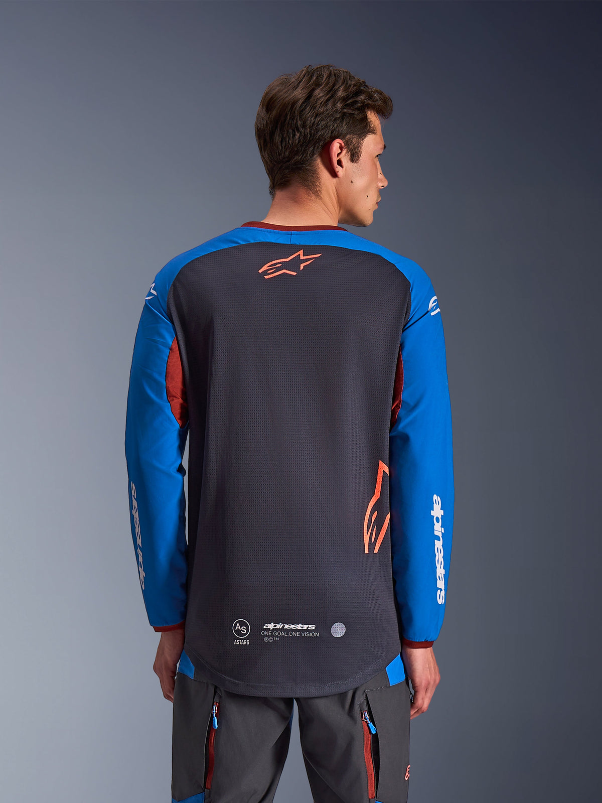 Alpinestars Maxdura Dual Jersey in Deep Gray and Royal Blue, back view of male model, mesh torso construction with orange Astars logo, long sleeves with white branding, maroon collar and cuffs, worn with gray technical pants, dual-sport and adventure motorcycle gear