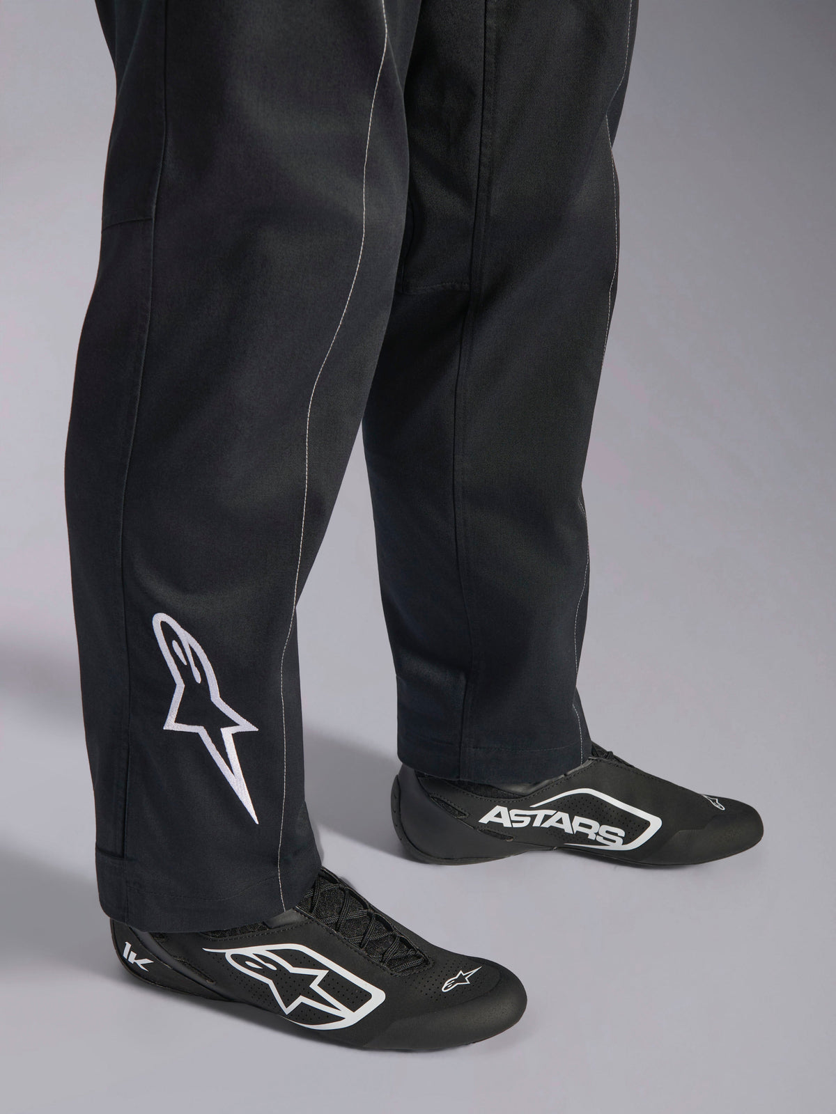 Alpinestars Kart Indoor V2 Suit, svart kartingoverall, närbild på underben och fötter med en vit Alpinestars-logotyp på vaden, bärs med matchande svartvita racingskor, designad för rekreationskarting