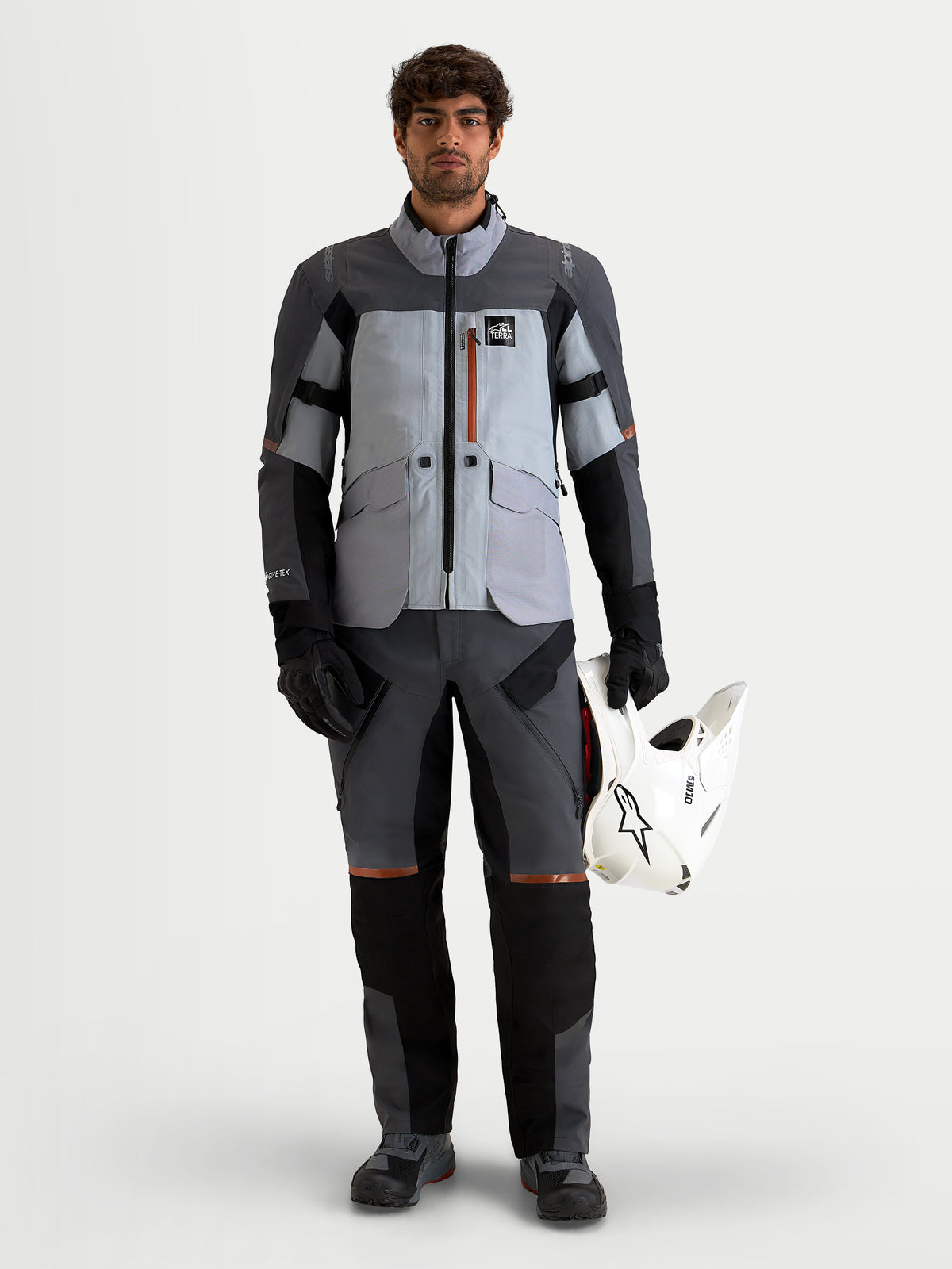 Alpinestars St-7 V2 2L Gore-Tex Gear Set, adventure touring motorcycle jacket and pants in light gray, dark gray, and black, worn by a model holding a white helmet, waterproof textile construction.