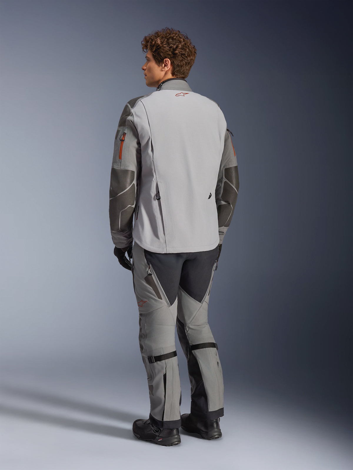 Alpinestars Nazca 3L GORE-TEX PRO-jacka, äventyrsmotorcykeljacka, mörkgrå och mellangrå, vy bakifrån bärs med matchande äventyrsbyxor och svarta stövlar, med mörkgrå Superfabric-förstärkningar på ärmarna, orangea dragkedjedetaljer och en liten röd logotyp på kragen bak.
