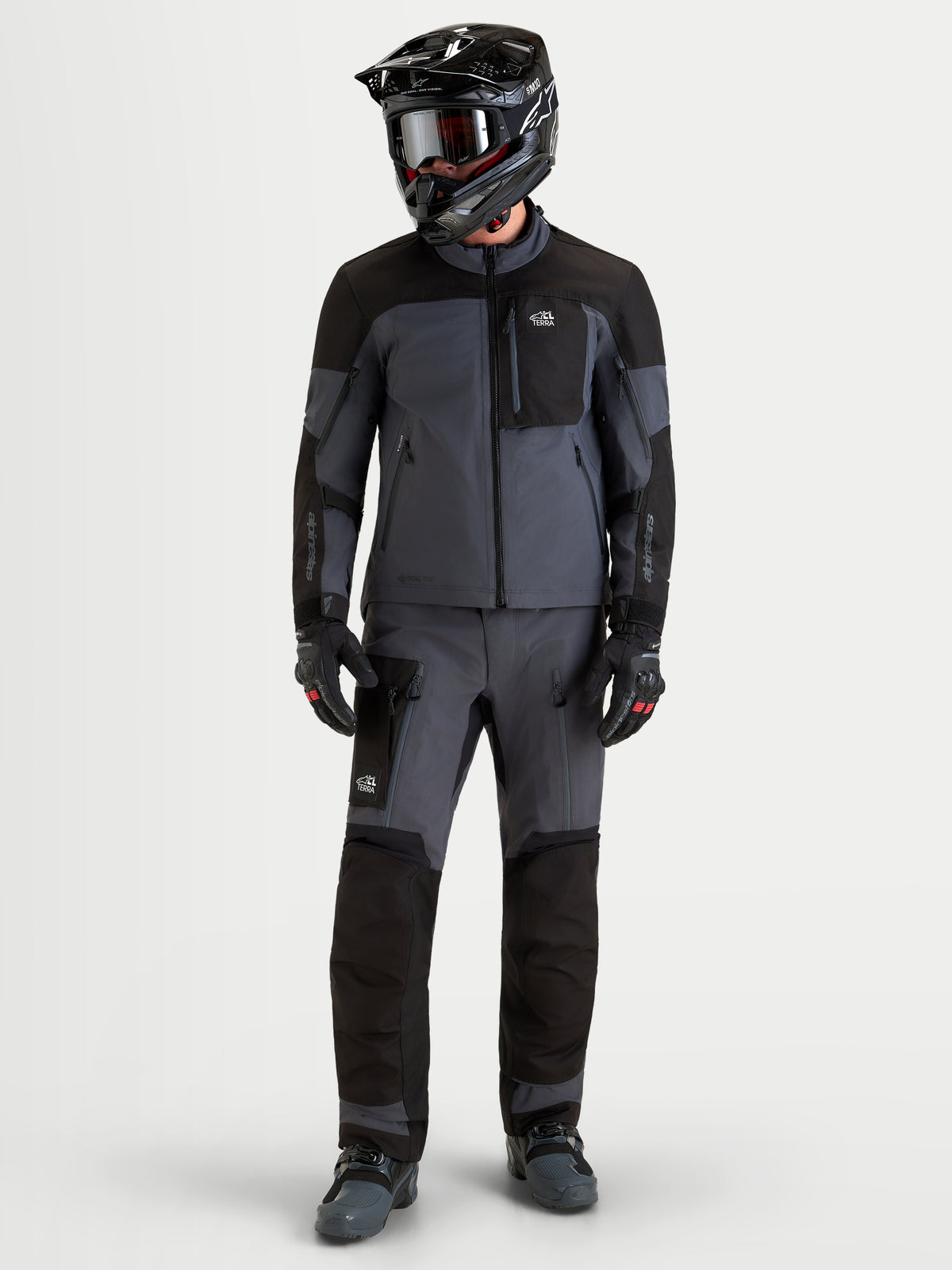 Alpinestars Arenis Gore-Tex jacka, äventyrsmotorcykeljacka för herr i svart och mörkgrå, bars av en förare med matchande hjälm och skyddsutrustning, framifrån i helkroppsvy