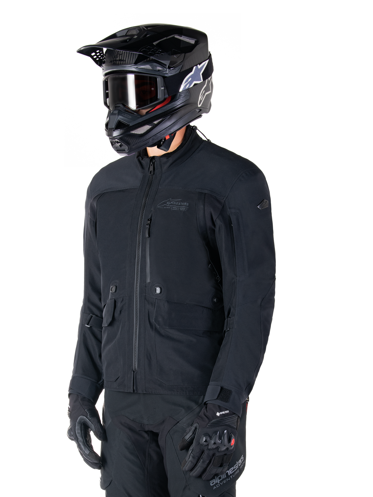 Alpinestars St-7 Prime GORE-TEX-jacka, svart motorcykeljacka, bärs av en förare med svart hjälm och goggles, stora bröstfickor i cargostil och ett laminerat vattentätt skal, premiumutrustning för adventure touring och pendling