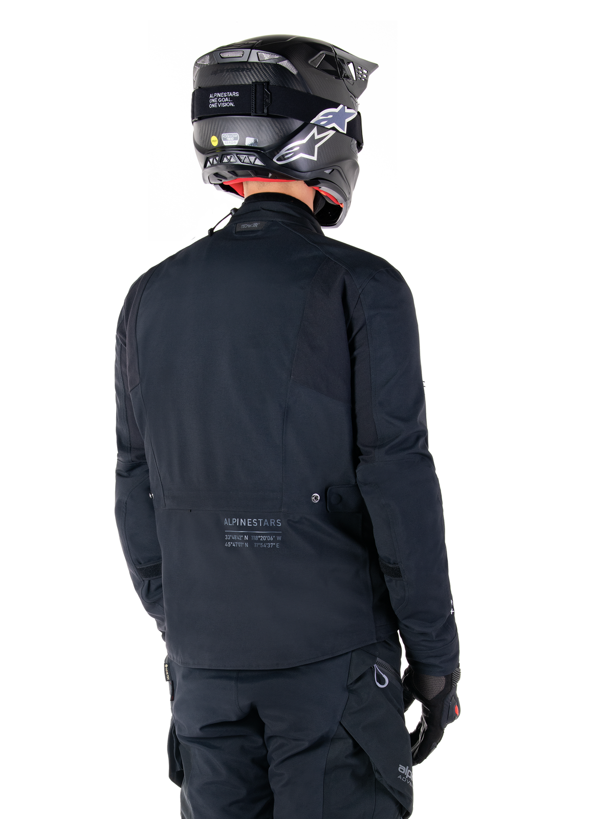 Alpinestars St-7 Prime GORE-TEX-jacka, svart, vy bakifrån på en förare som bär jackan med matchande hjälm och byxor, med Alpinestars-logotyp och geografiska koordinater på nedre ryggen.