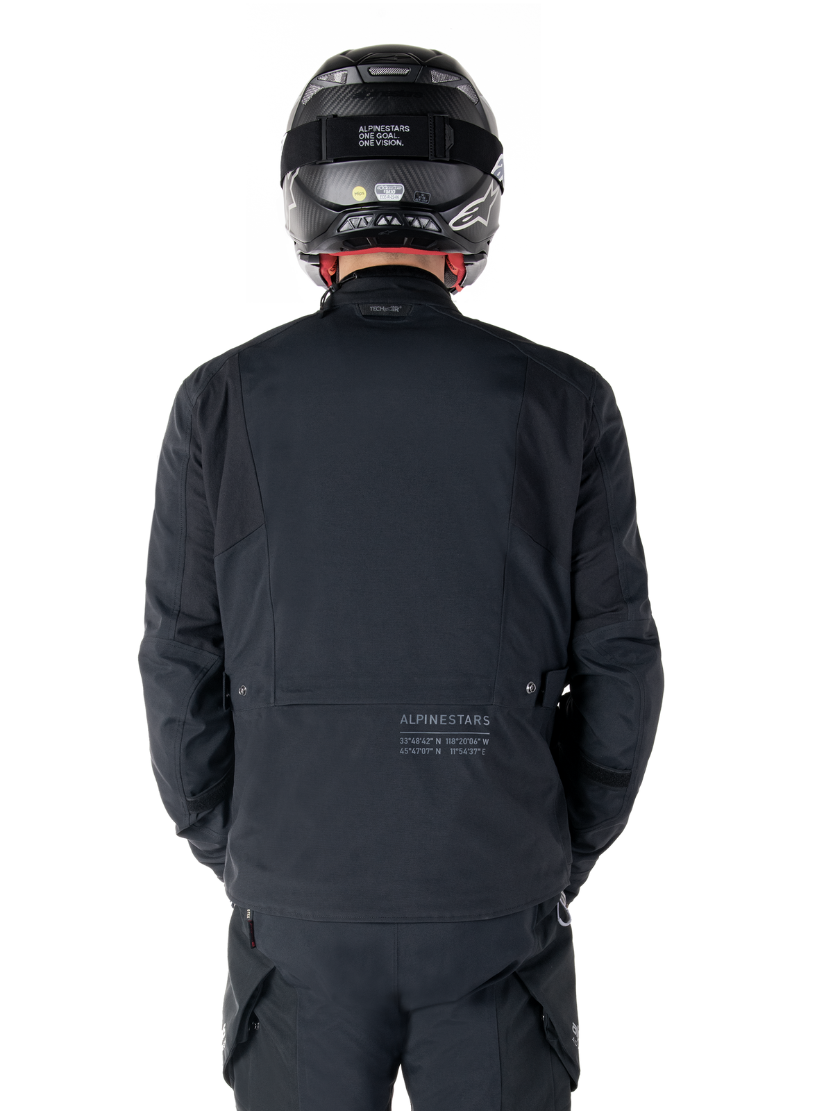 Alpinestars St-7 Prime GORE-TEX-jacka, svart, vy bakifrån av en förare med subtil logotyp och geografiska koordinater på nedre delen av ryggen, bärs med matchande svart hjälm och byxor, stilren och teknisk motorcykelutrustning