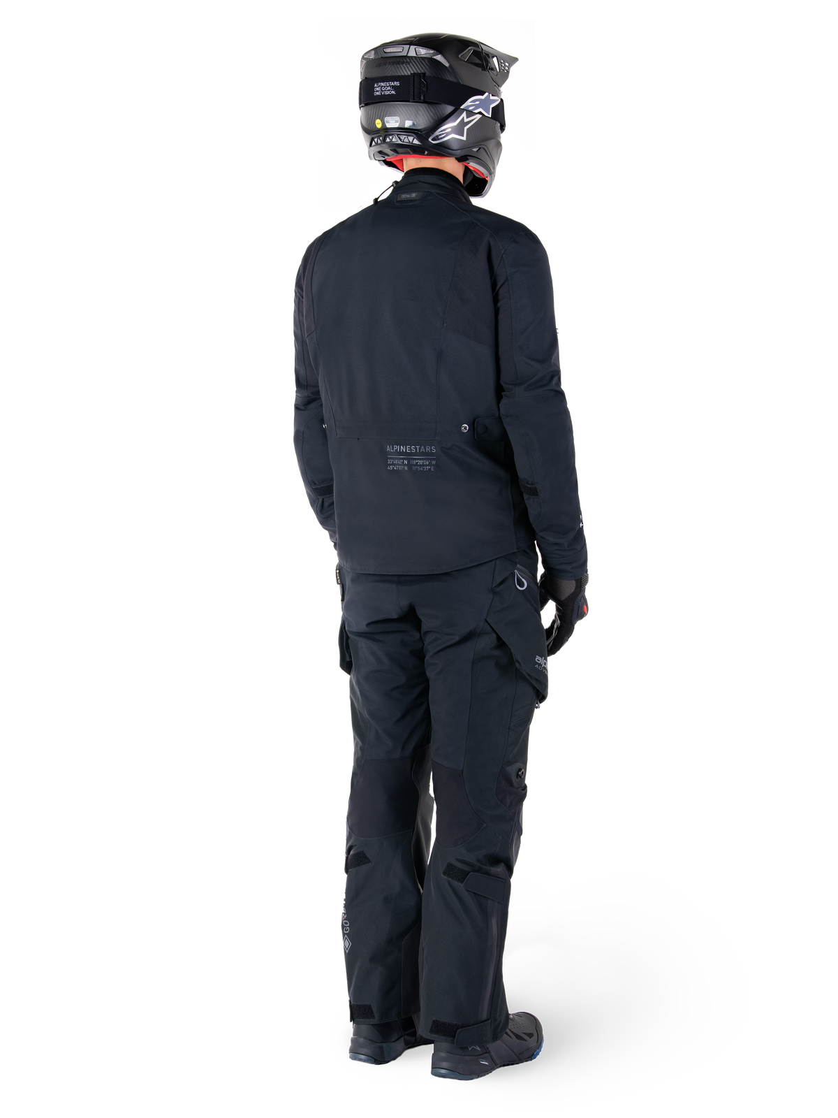 Vy bakifrån av en förare som bär Alpinestars St-7 Prime GORE-TEX-jacka i svart, kombinerat med matchande svarta touringbyxor, en svart motorcykelhjälm med vit logotyp och svarta körstövlar, komplett skyddande adventure-utrustning för touring och pendling.