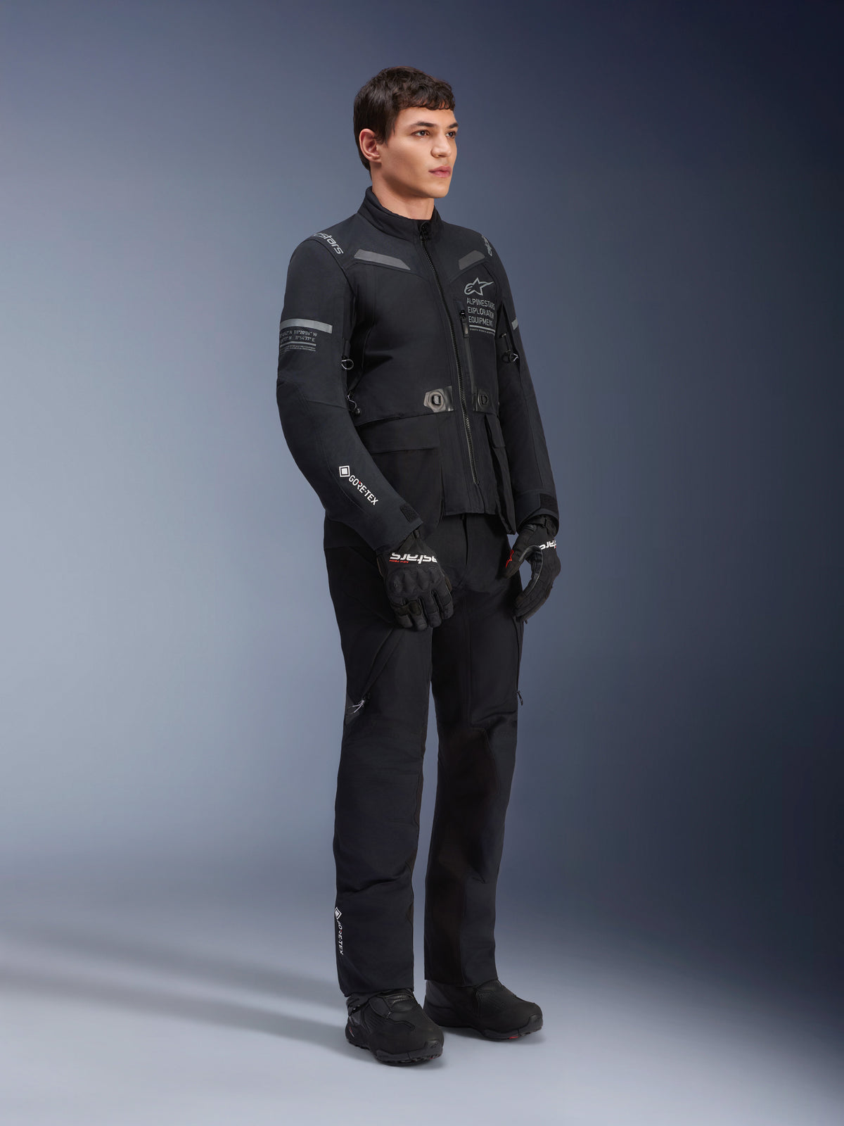 Manlig modell iförd Alpinestars ST-7 2L GORE-TEX-jacka i Black Dark Gray, motorcykeljacka för äventyr och touring, matchande svarta byxor och handskar, trekvarts helkroppsvy, med GORE-TEX-logotyp och Alpinestars Exploration Equipment-märkning