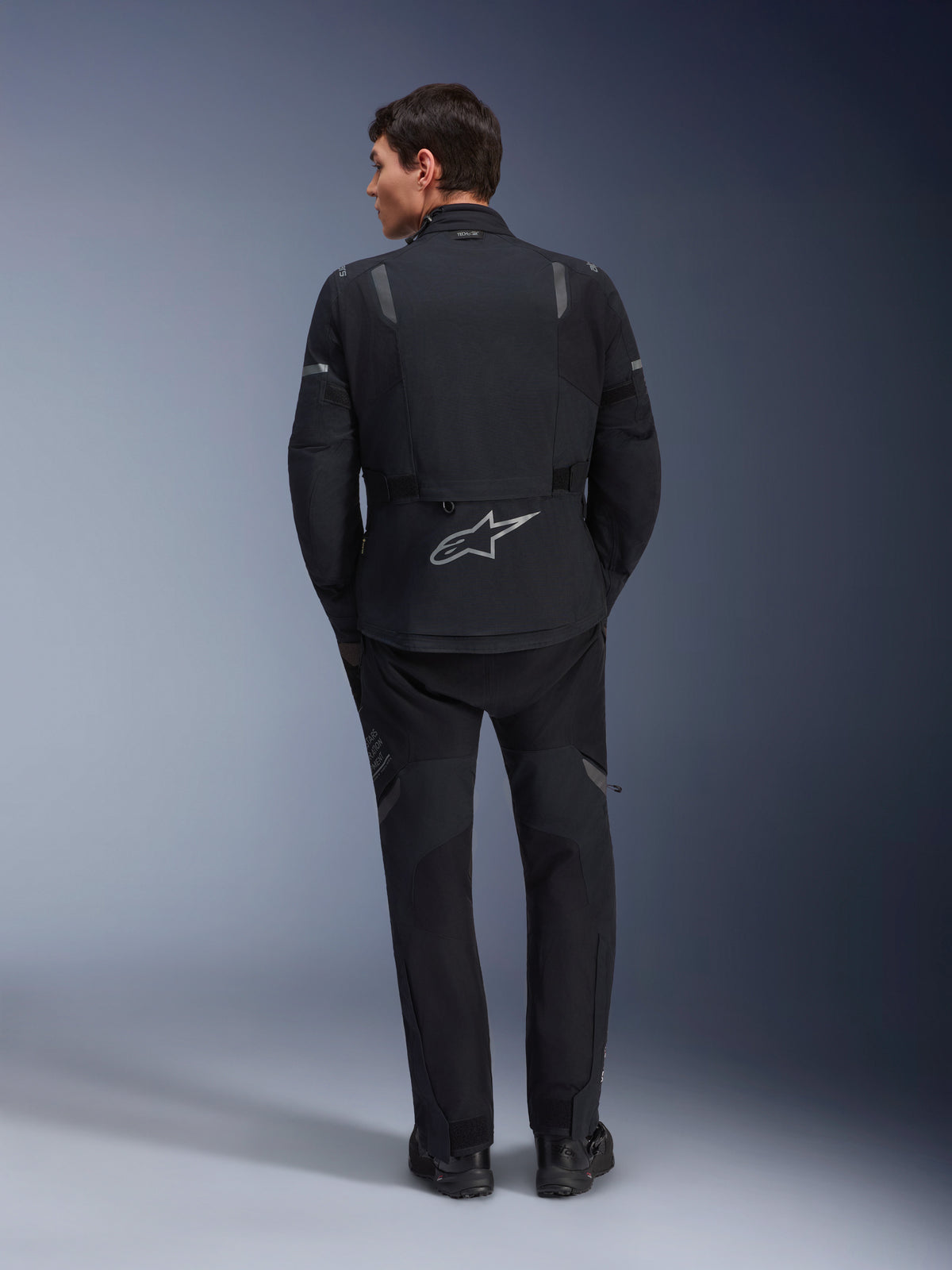 Alpinestars ST-7 2L GORE-TEX-jacka och matchande ST-7 2L GORE-TEX-byxor, svarta och mörkgrå äventyrsmotorcykelkläder, vy bakifrån som visar reflekterande Alpinestars-logotyp på jackan och teknisk textilkonstruktion.