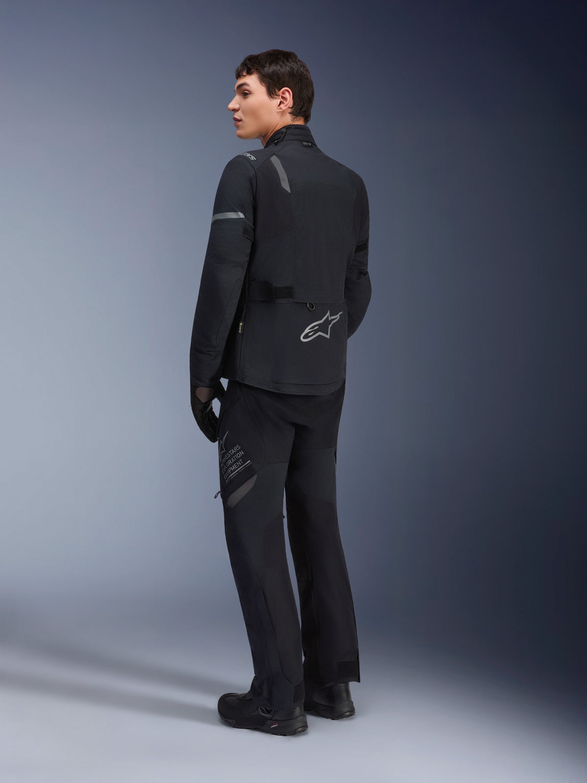 Manlig modell bär Alpinestars ST-7 2L GORE-TEX-jacka i Black Dark Gray, sedd snett bakifrån med reflekterande detaljer och silverlogotyp på ländryggen, adventure touring-kläder för motorcykel, kombinerat med matchande svarta körbyxor och stövlar