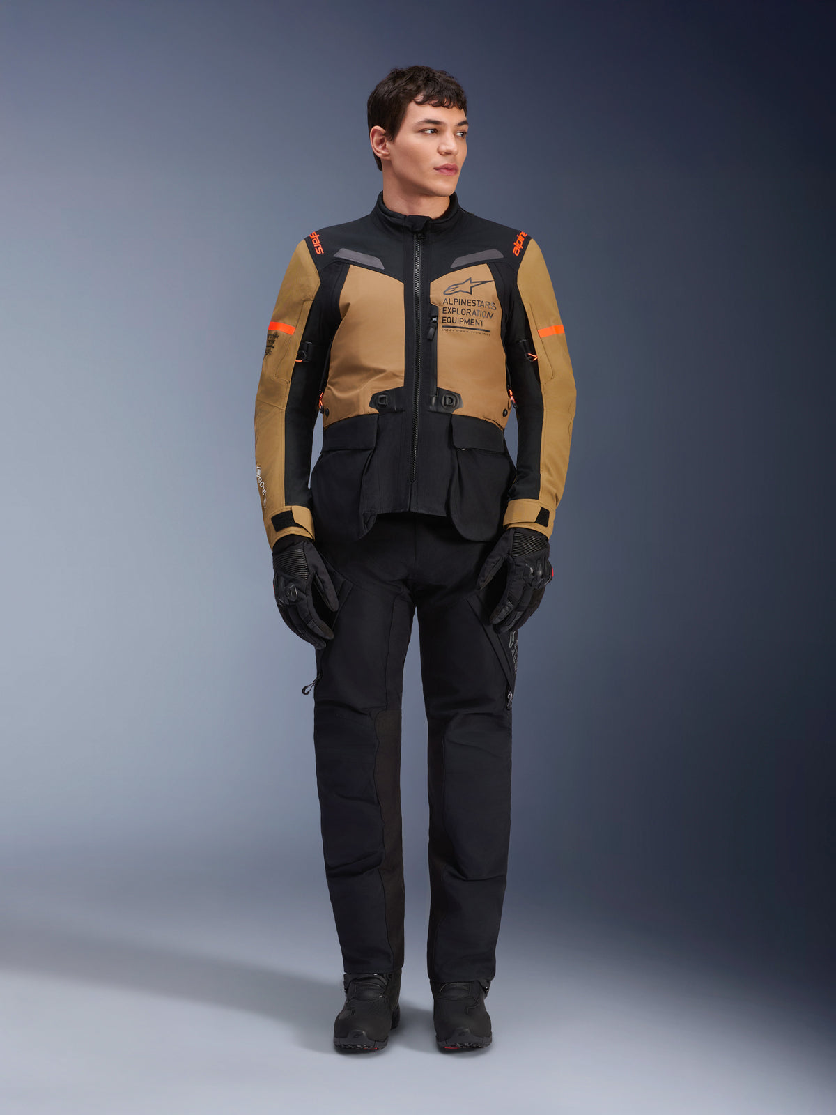 Manlig modell som bär Alpinestars ST-7 2L GORE-TEX-jacka, Gobi Brown och svart, äventyrsmotorcykelutrustning med matchande svarta byxor och handskar, med Alpinestars Exploration Equipment-logotyp på bröstet, funktionell vattentät design för touring på landsväg och i terräng