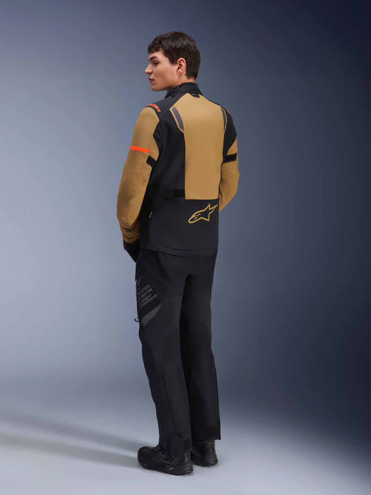 Alpinestars ST-7 2L GORE-TEX-jacka, äventyrsmotorcykeljacka, Gobi Brown och svart, sedd bakifrån på en modell som bär matchande mc-byxor och stövlar, med orangea detaljer på ärmarna, vattentätt laminerat skal och Alpinestars-logotyp, designad för touring och vägkörning