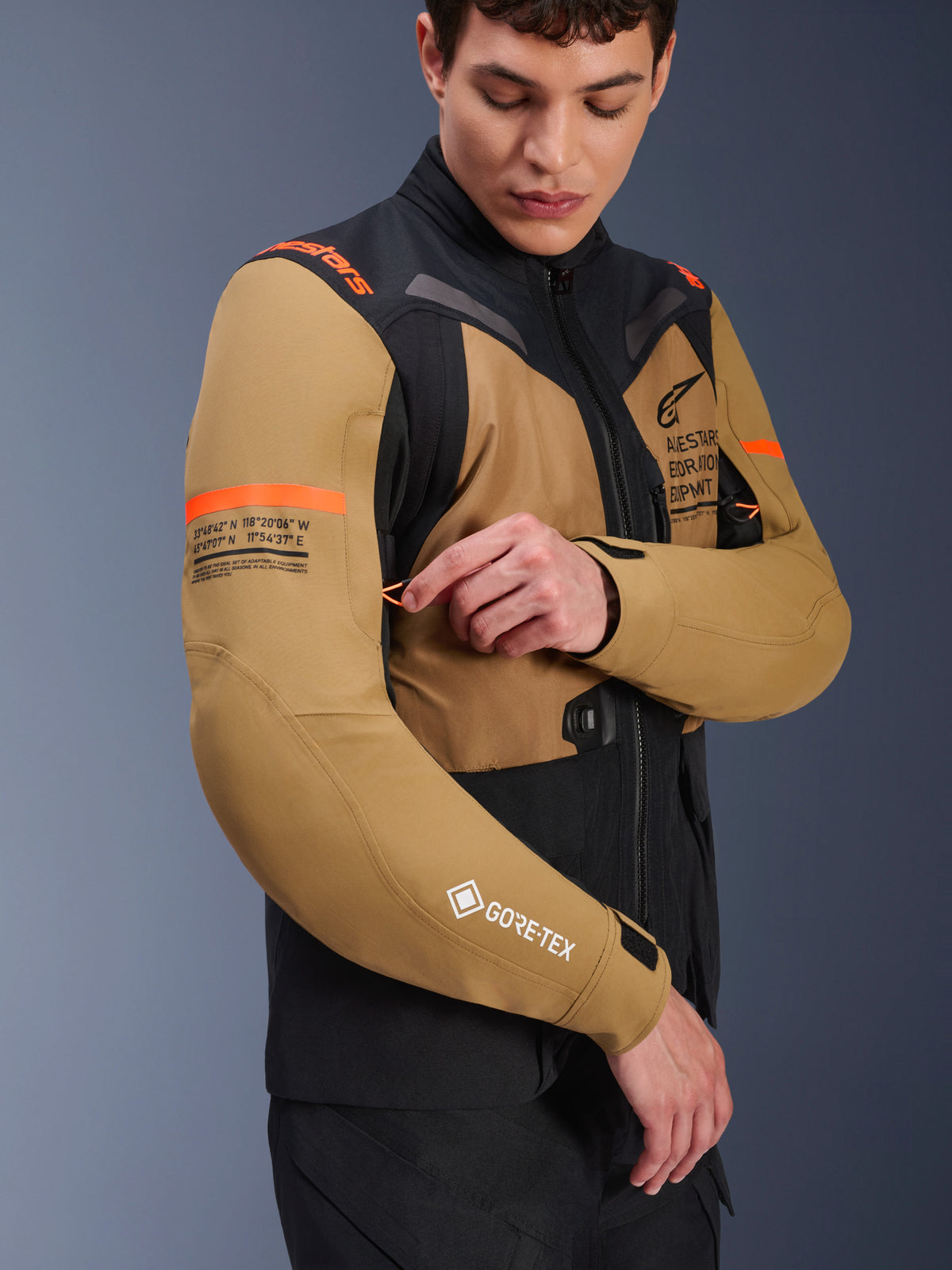 Alpinestars ST-7 2L GORE-TEX-jacka i Gobi Brown och svart, adventure-touring motorcykeljacka buren av en modell som justerar en ventilationsöppning på ärmen, med orangea detaljer, koordinatgrafik och GORE-TEX-logotyp