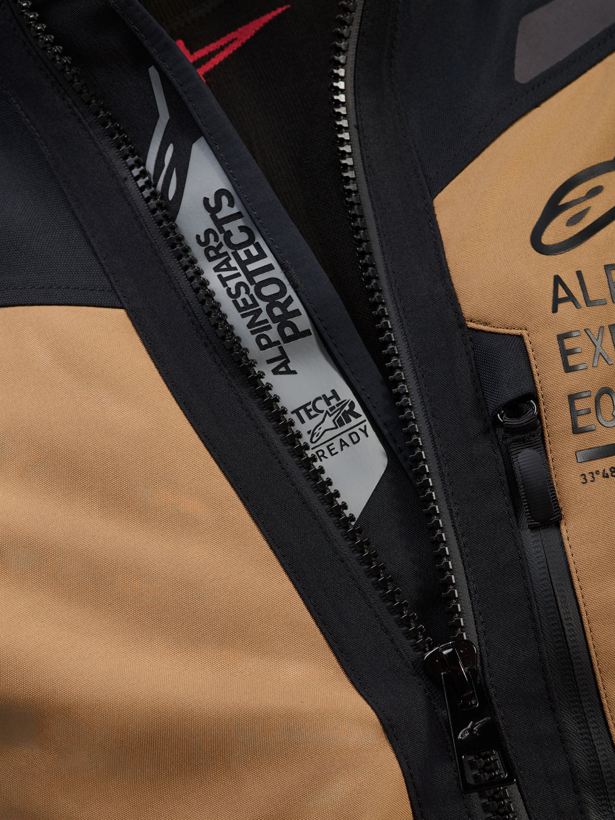 Närbild på Alpinestars ST-7 2L GORE-TEX-jacka i Gobi Brown och svart, med 'ALPINESTARS PROTECTS' och 'TECH-AIR READY'-märkning innanför huvuddragkedjan, robust textilkonstruktion och logotyper på bröstet