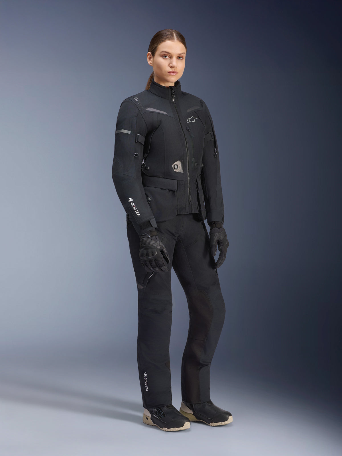 Kvinnlig modell bär Alpinestars Stella ST-7 2L GORE-TEX adventure touring-ställ, svart och mörkgrå jacka och byxor, med tydlig GORE-TEX-branding på ärmen och benet, kombinerat med svarta handskar och MC-stövlar, professionell motorcykelutrustning designad för touring i alla väder.