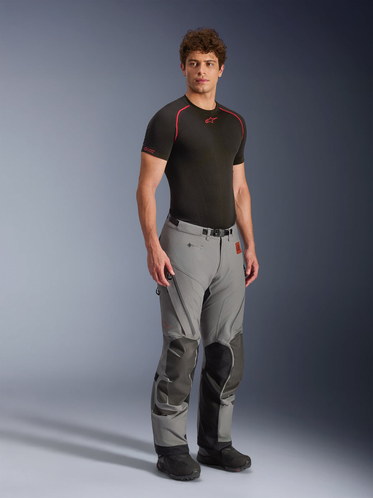 Male model wearing Alpinestars Nazca 3L GORE-TEX PRO Pants, Dark Gray and Black adventure motorcycle pants, featuring reinforced knee panels and zippered ventilation, worn with a black base layer top, technical gear designed for extreme all-terrain riding.