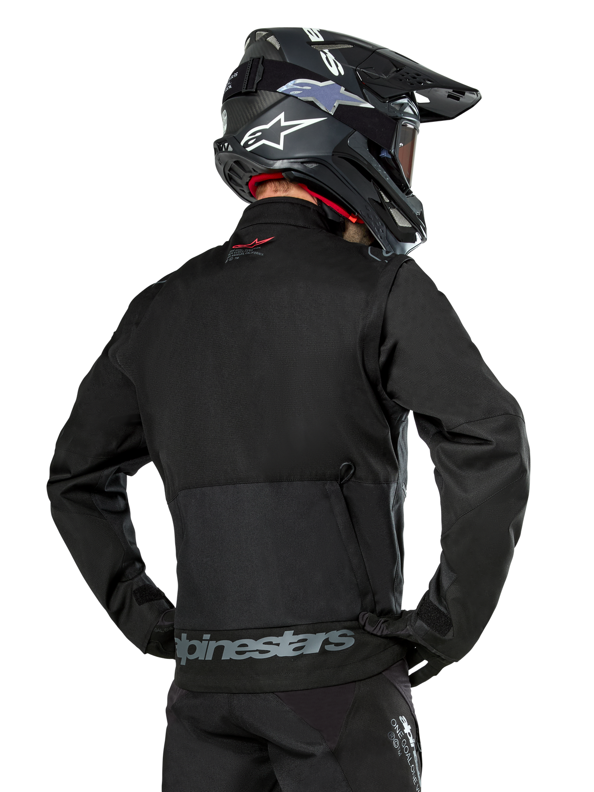 Ryggvy av en förare som bär Alpinestars Pro-Dura Jacket i svart, slitstark endurojacka i ripstop-textil med en stor bakre förvaringsficka, grå Alpinestars-logotyp över nedre delen av ryggen, tillsammans med en svart motocrosshjälm.