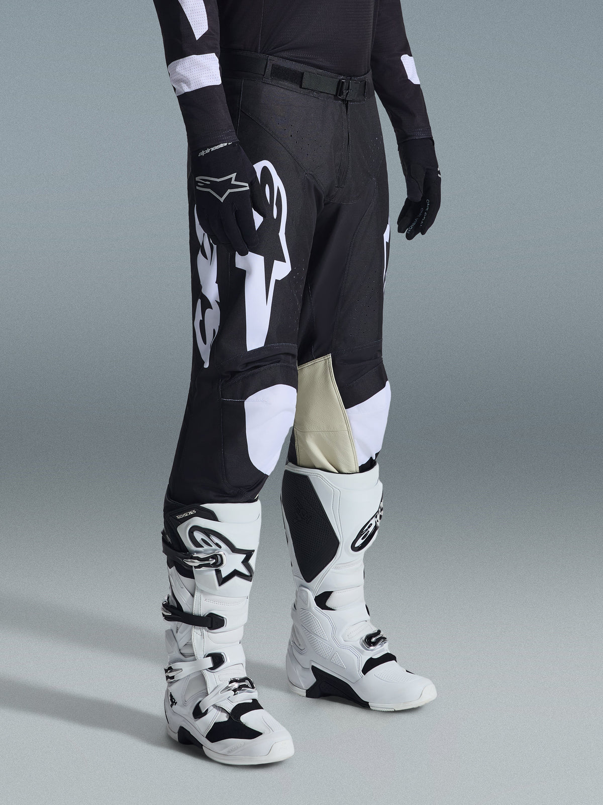 Alpinestars 2026 Racer Air Riway Pants, Black and White motocross pants with large white logo graphics, beige leather inner knee panels, worn with white and black off-road boots and black gloves in a studio setting.
