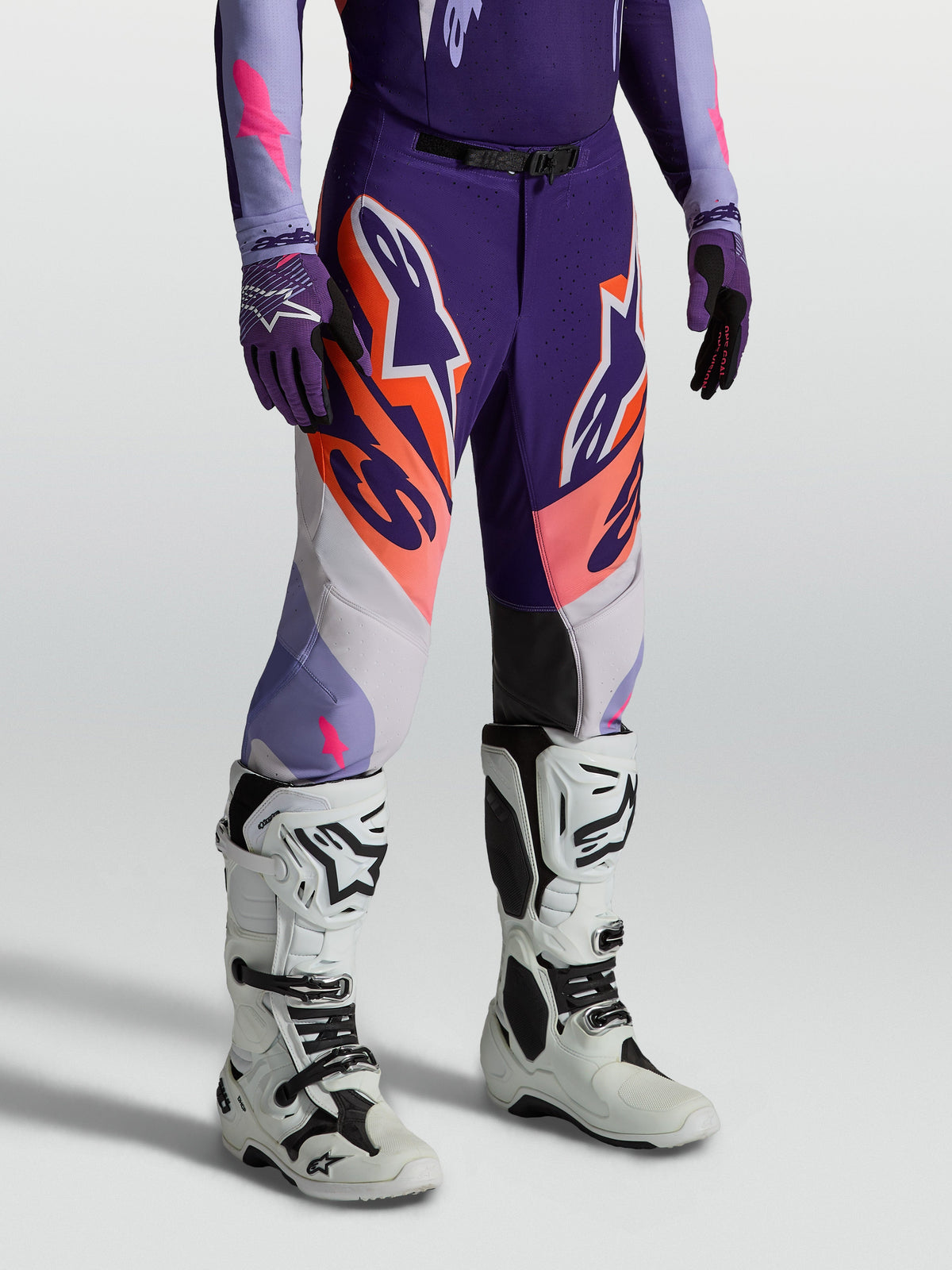 Alpinestars Supertech Dyna byxor, motocrossbyxor i lila, rosa fluo och orange, atletisk tävlingspassform, bärs med vita stövlar för professionell terrängkörning