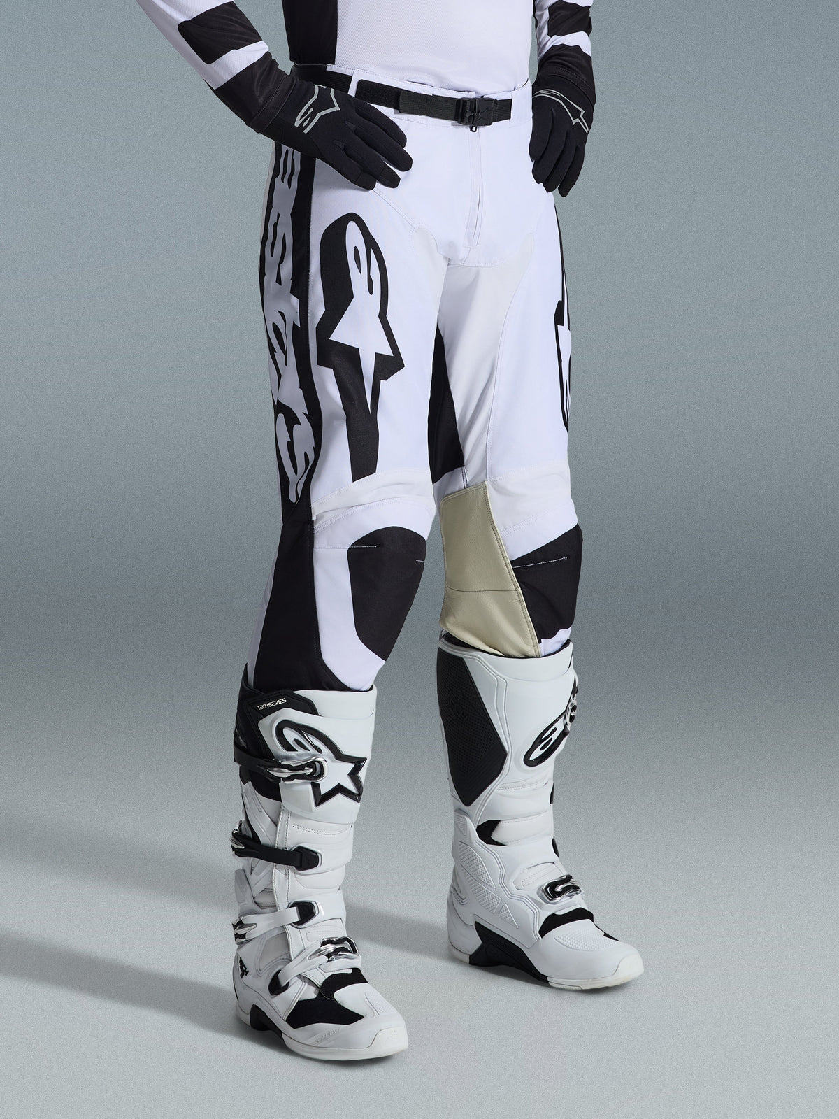 Alpinestars 2026 Racer Riway-byxor, motocrossbyxor, vita och svarta med framträdande svart Alpinestars-grafik och logotyper, beige läderförstärkningar på insidan av knäna, bärs med vita och svarta motocross-stövlar och svarta handskar, professionell racingutrustning för terrängkörning.