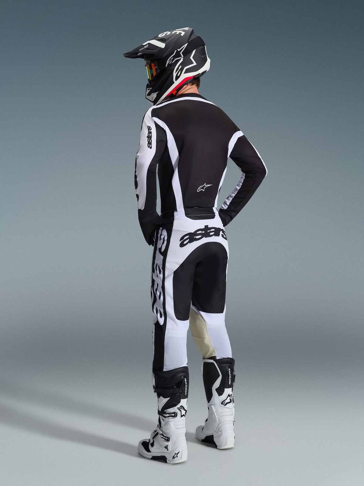 Motocrossförare sedd bakifrån klädd i Alpinestars 2026 Racer Riway-byxor i vitt och svart, matchande svart och vit tröja med tydlig logotyp, svart och vit hjälm och vita motocross-stövlar, komplett racingutrustning för terrängkörning.