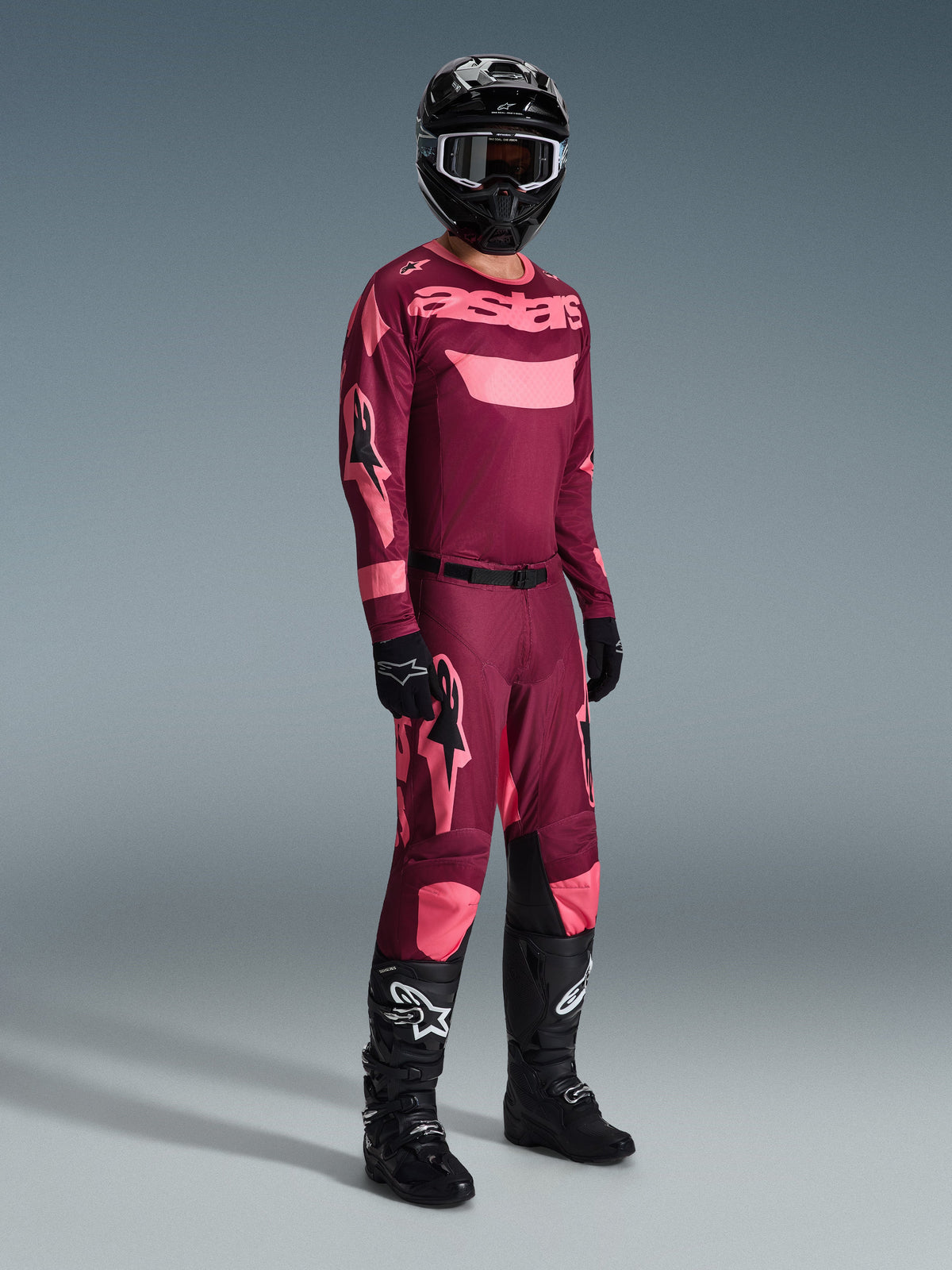 Motocrossförare i komplett racingutrustning, Alpinestars 2026 Racer Riway-tröja i Dark Red Coral med rosa detaljer, matchande mörkröda byxor med rosa logotyper, svart hjälm med vitkantade goggles, svarta handskar och svarta stövlar med vita logotyper, koordinerad outfit för terrängkörning