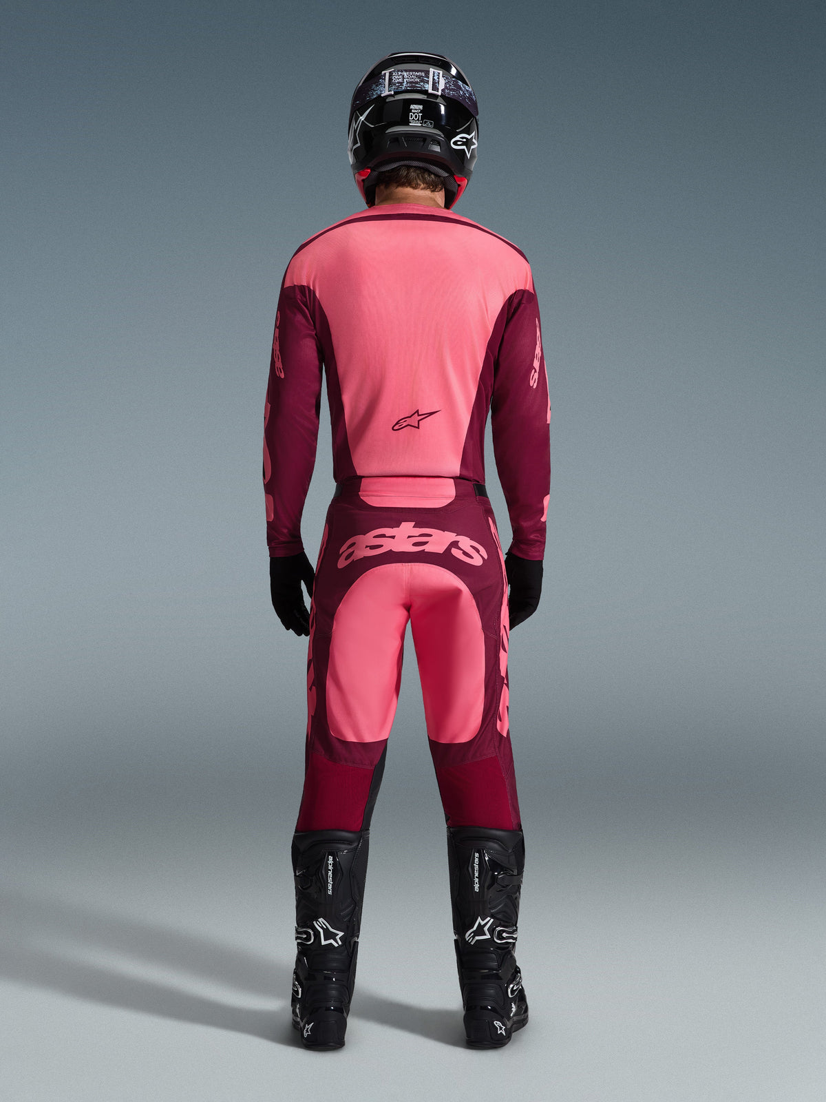 Motocrossförare sedd bakifrån klädd i Alpinestars 2026 Racer Riway-tröja och matchande byxor i Dark Red Coral, med svarta stövlar, en svart hjälm och svarta handskar, koordinerad högpresterande utrustning för terrängkörning