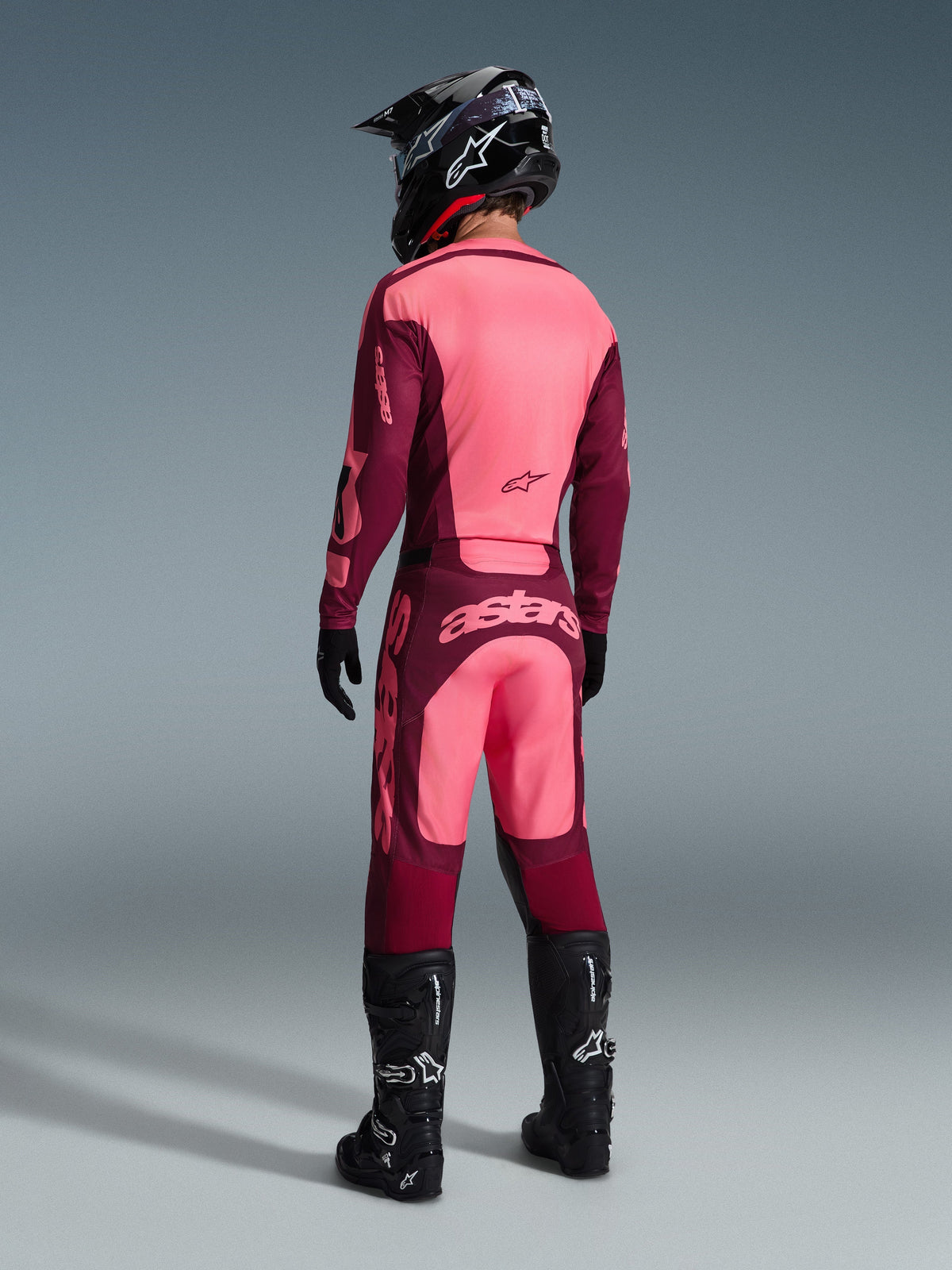 Alpinestars 2026 Racer Riway-tröja och matchande byxor, motocrossutrustning i Dark Red Coral, vy bakifrån av förare med svart hjälm och svarta stövlar, livfulla korallfärgade och vinröda paneler med tydlig Astars-logotyp på ryggen