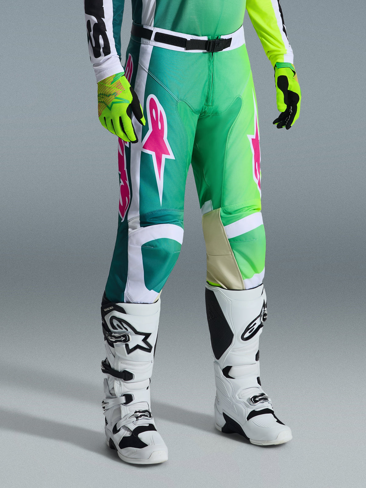 Alpinestars 2026 Racer Portl byxor, motocrossbyxor, gröna, vita och svarta med rosa logotyper, bärs med vita stövlar och neongula handskar, med djärva grafiska mönster och ett justerbart svart midjespänne, prestandautrustning för terrängkörning.
