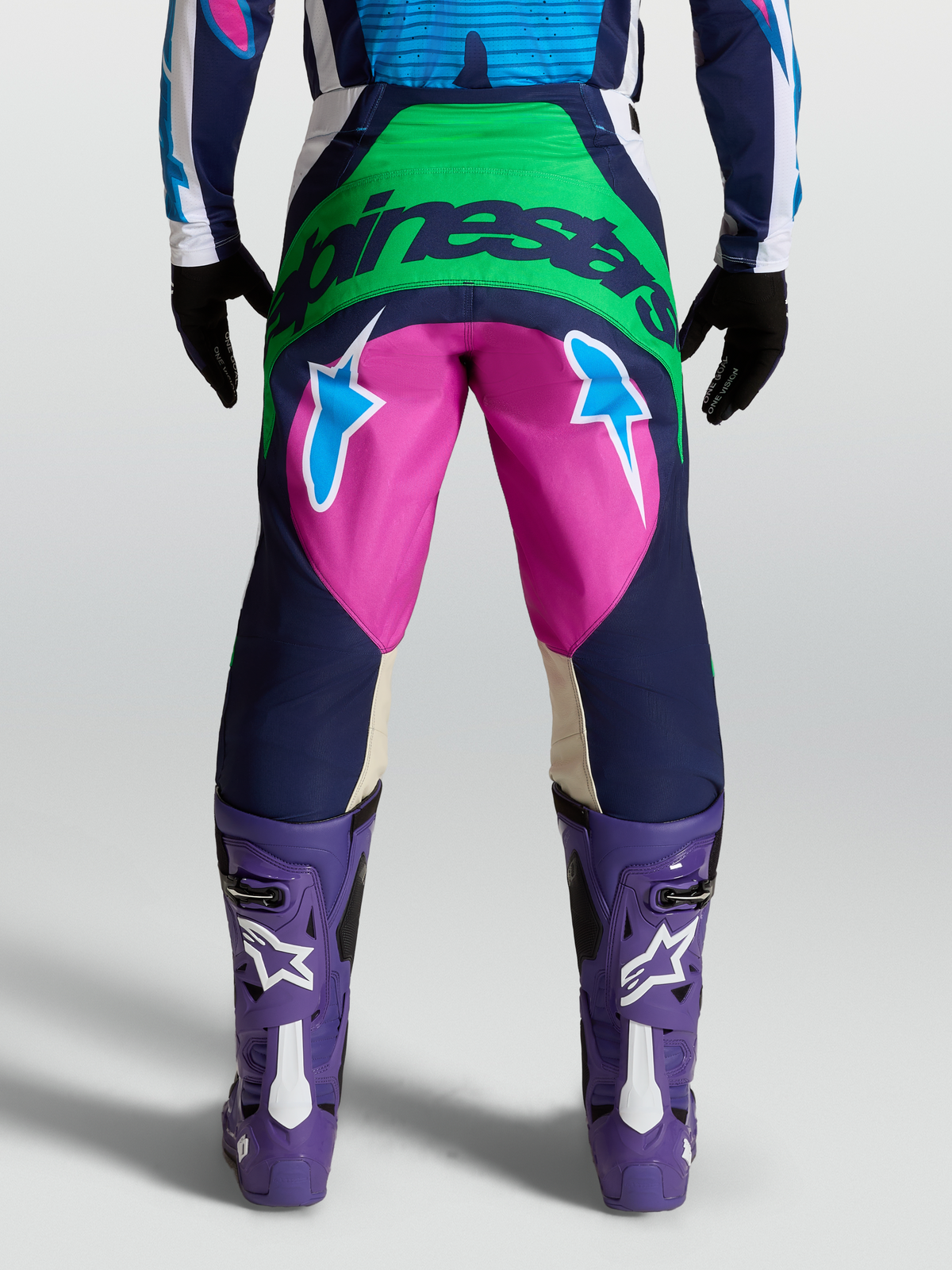 Alpinestars Techstar Maven byxor, blå och rosa motocrossbyxor på modell i vy bakifrån, gröna och blå stjärngrafiker, förarskydd för terrängkörning.
