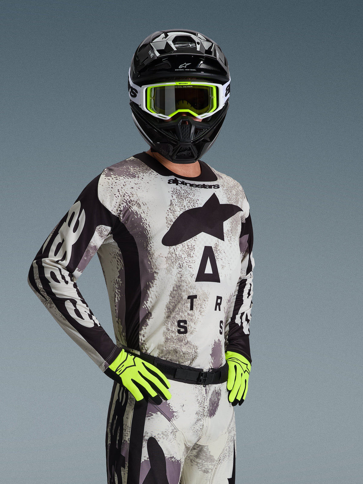 Motocross rider wearing the Alpinestars 2026 Racer Lahnd Jersey and gear set in Sand Camo, light gray camouflage pattern with black logo branding, black helmet with white and neon yellow goggles, bright neon yellow gloves, professional off-road racing apparel