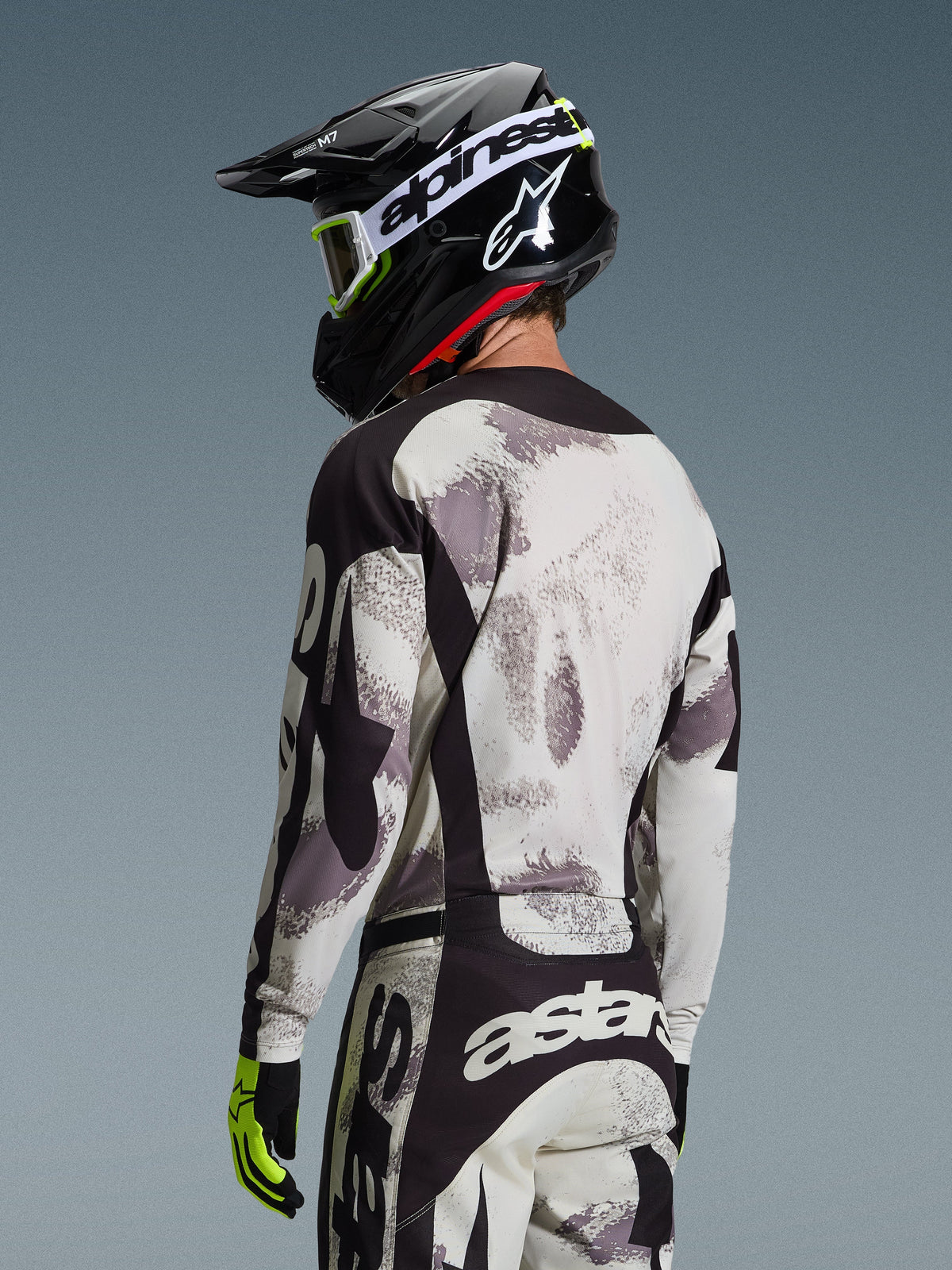 Motocross rider from the back wearing the Alpinestars 2026 Racer Lahnd Gear Set, Gray and White Sand Camo jersey and pants with black accents, black helmet with white Alpinestars logo, neon yellow and black gloves, coordinated off-road performance outfit