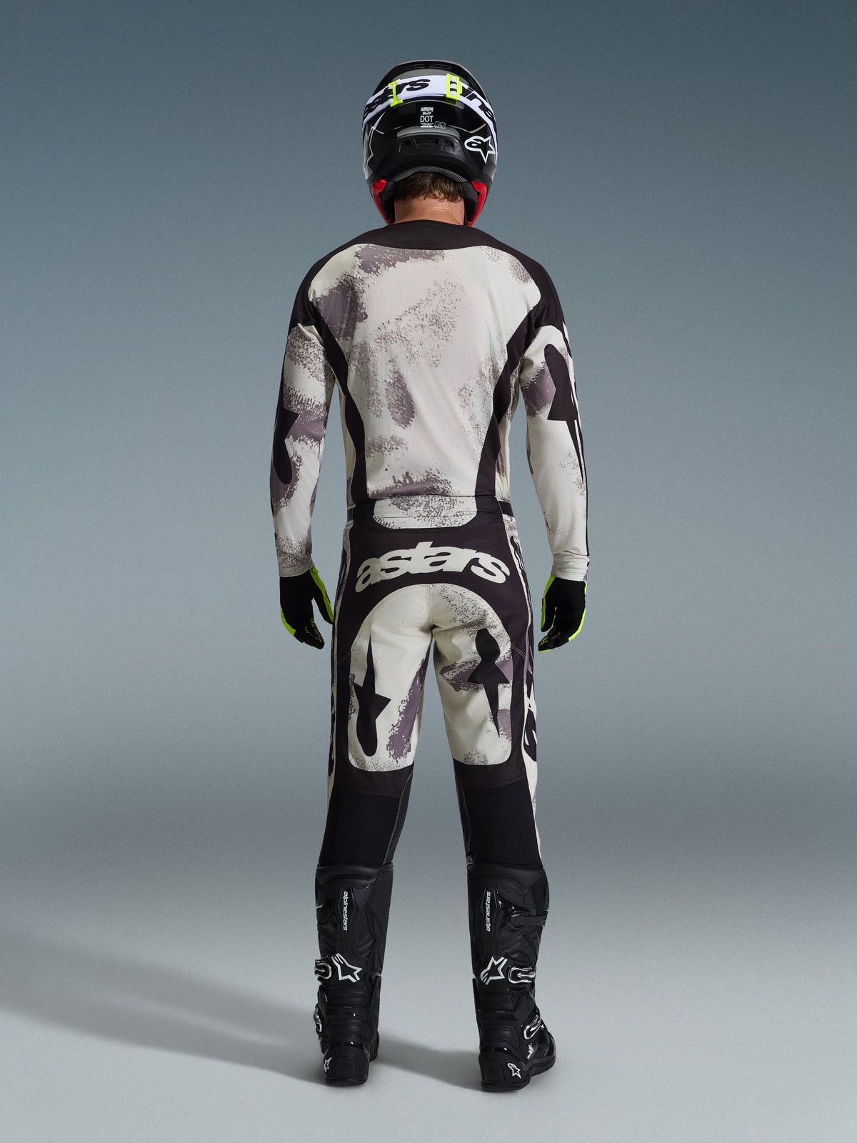 Motocross rider wearing complete Alpinestars 2026 Racer Lahnd Gear Set, viewed from behind, Sand Camo jersey and pants with light gray and black abstract pattern, black helmet, black and neon yellow gloves, black boots, featuring 'astars' branding on the rear of the pants
