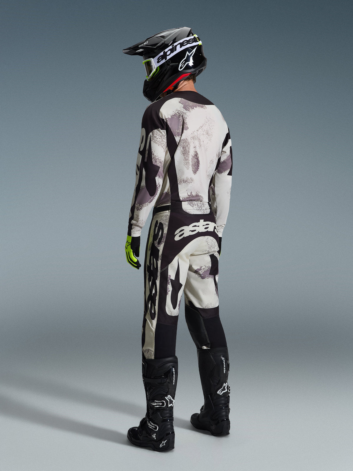 Motocross rider wearing Alpinestars 2026 Racer Lahnd Gear Set, Sand Camo abstract pattern in gray and white, black pants with bold white Alpinestars branding, black helmet and boots, fluorescent green accents on gloves, complete outfit for off-road racing