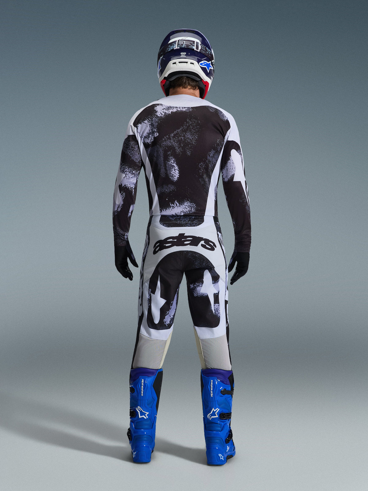 Motocross rider from behind wearing the Alpinestars 2026 Racer Lahnd Jersey and matching pants in Iron Camo, featuring a black and white splatter pattern, paired with bright blue boots and a coordinating helmet, complete gear set for off-road racing