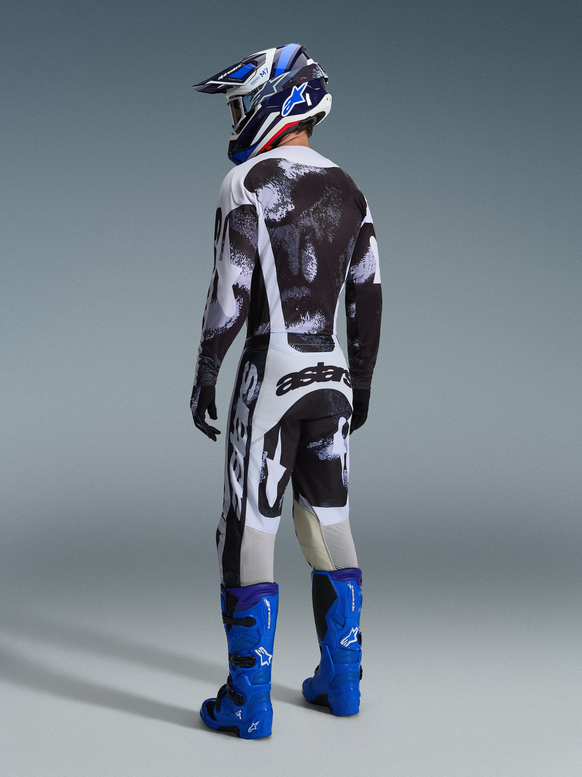 Motocross rider wearing complete racing gear from the back, 2026 Racer Lahnd Jersey and pants in Iron Camo, black grey and white abstract pattern, bright blue Alpinestars boots, blue and white helmet, coordinated outfit for off-road racing
