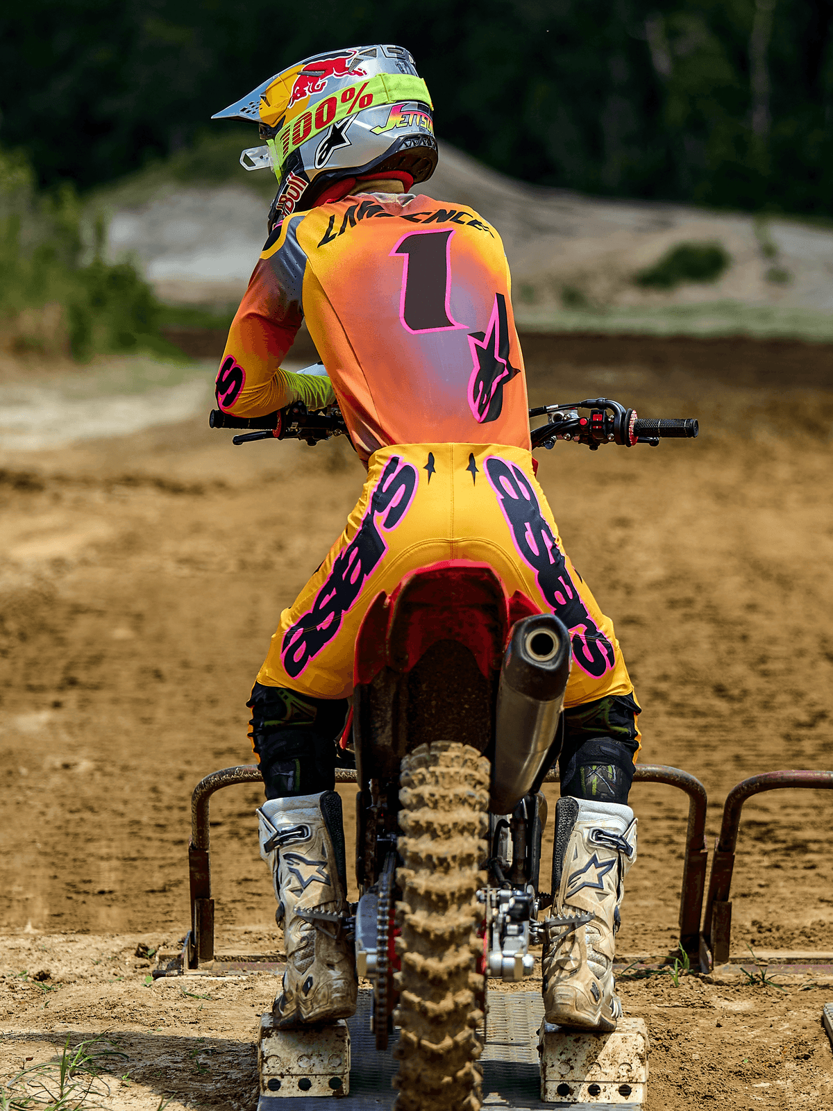 Motocrossförare sedd bakifrån bär Alpinestars 2025 Supertech Lipan-utrustningsset i gult och rosa, tröja med nummer 1 och 'LAWRENCE' på ryggen, gula byxor med rosa 'ASTARS'-branding, vita stövlar och matchande hjälm, sittande på en motocrosscykel vid startgrinden