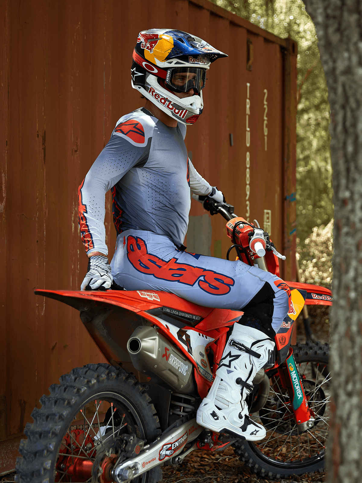 Motocrossförare på en röd motocrosscykel, klädd i Alpinestars 2025 Supertech Lipan utrustningsset i Gray Orange Fluo, ljusblå byxor med stor orange Astars-logotyp, matchande tröja, vita motocrossstövlar och en Red Bull-hjälm, koordinerad professionell racingutrustning