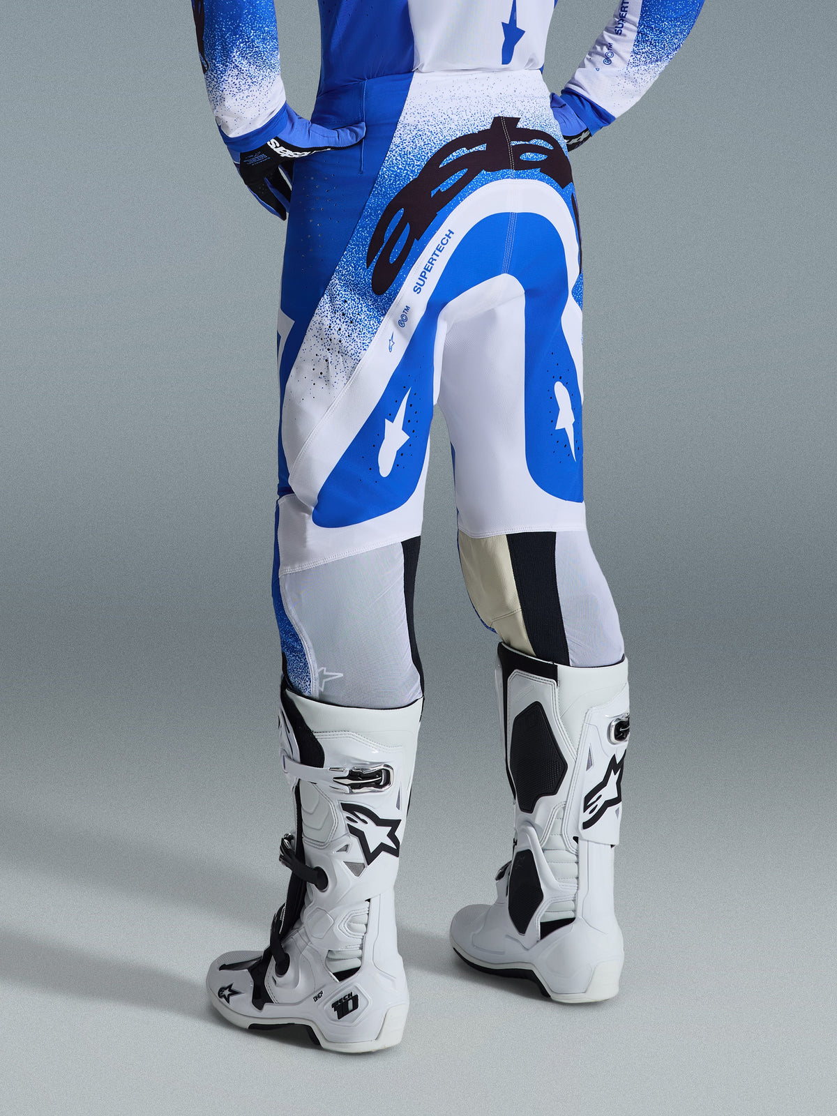 Alpinestars 2026 Supertech Scenz-byxor, motocrossbyxor, Ucla-blå och vita, vy bakifrån som visar en stor svart Astars-logotyp på sätet och 'SUPERTECH'-märkning, bärs med vita Tech 10-stövlar och en matchande blå och vit tröja.