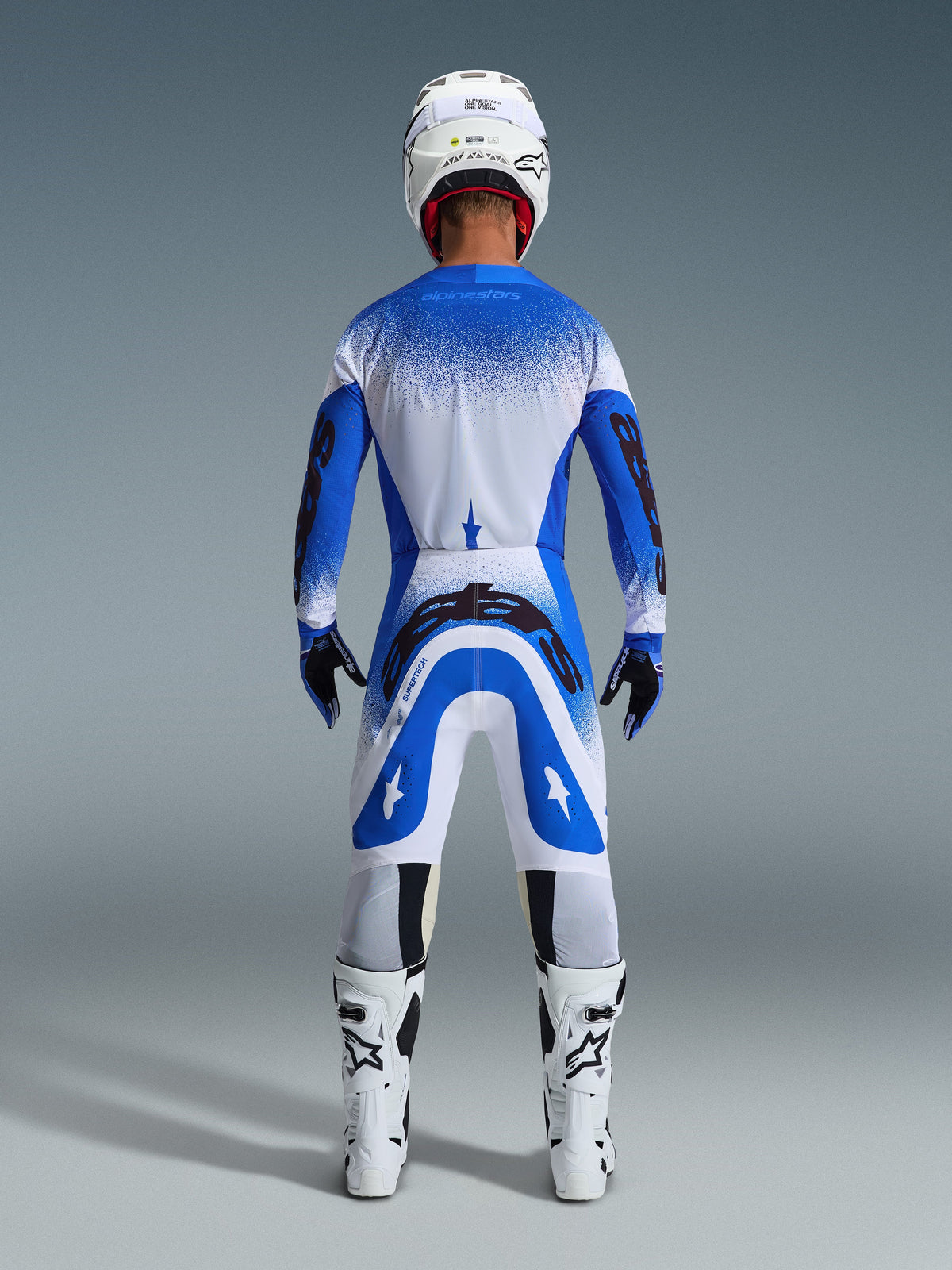Motocrossförare sedd bakifrån i full racingutrustning, Alpinestars 2026 Supertech Scenz-byxor i Ucla Blue och vitt, matchande blå och vit tröja med spräckligt mönster, vit hjälm, vita och svarta stövlar, koordinerad outfit för professionell racing
